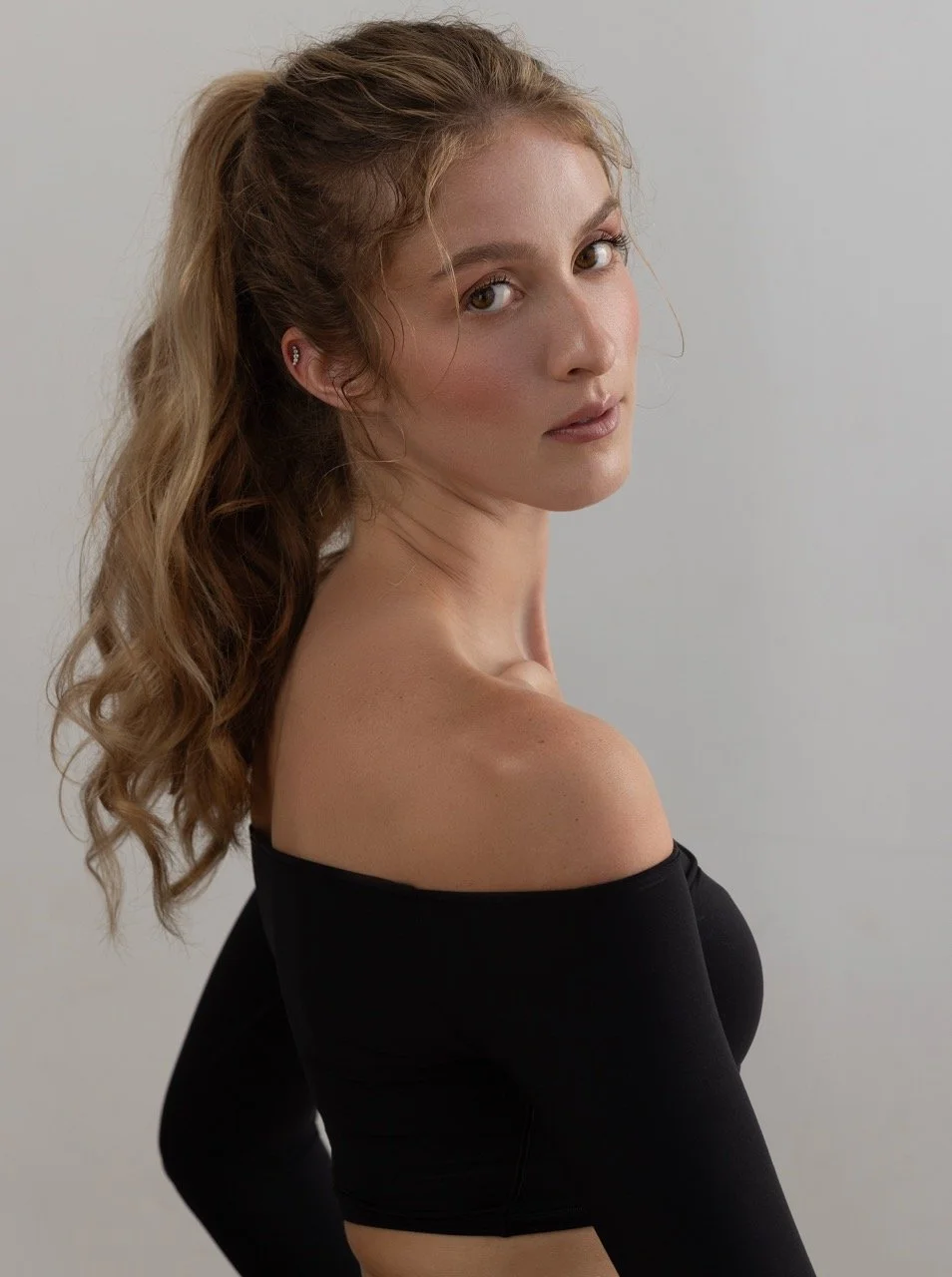A young woman with long, wavy blonde hair and fair skin, wearing an off-the-shoulder black top, looks over her shoulder at the camera against a plain gray background.