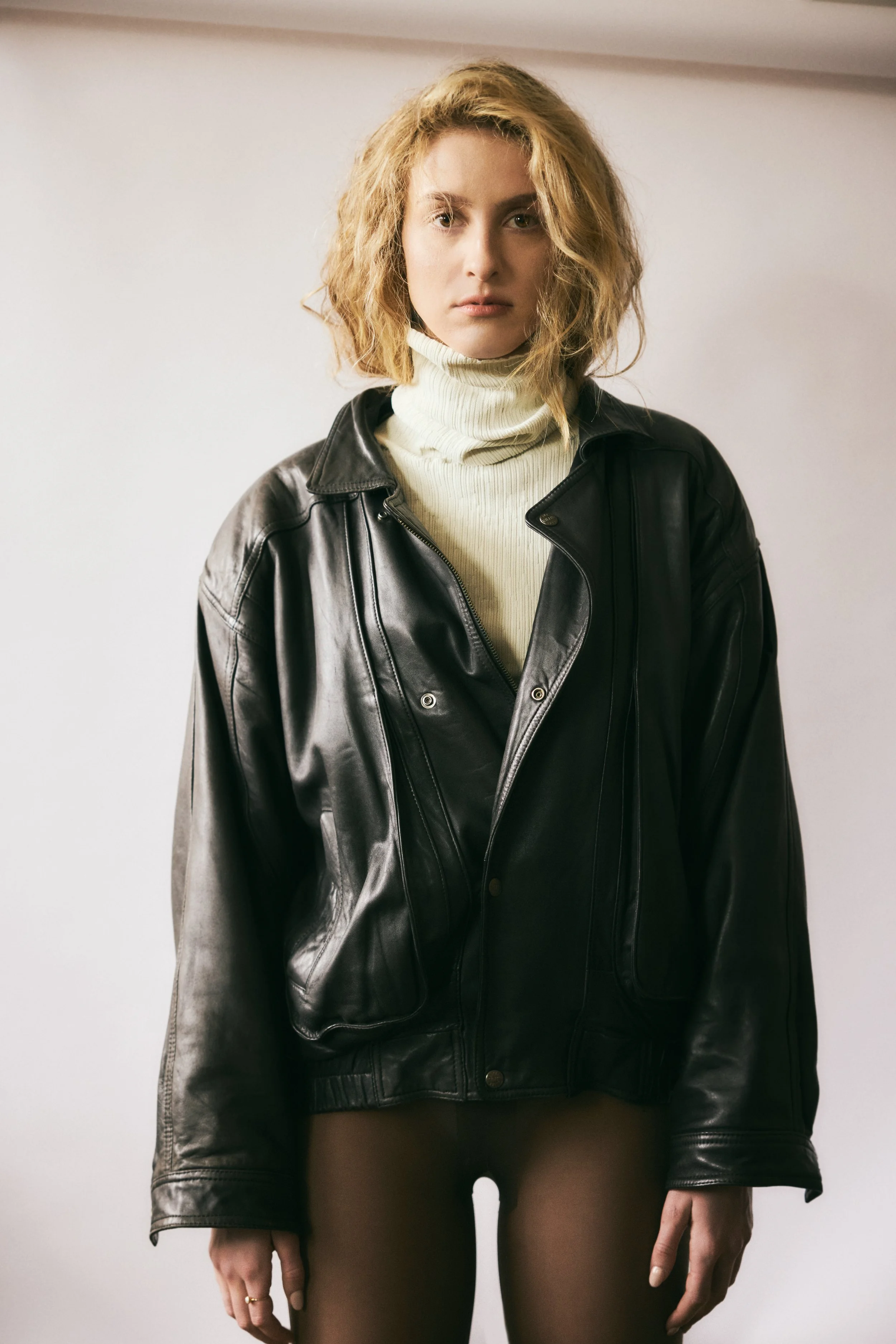 A young woman with curly blonde hair wearing a black leather jacket over a cream turtleneck sweater, standing against a plain white background.
