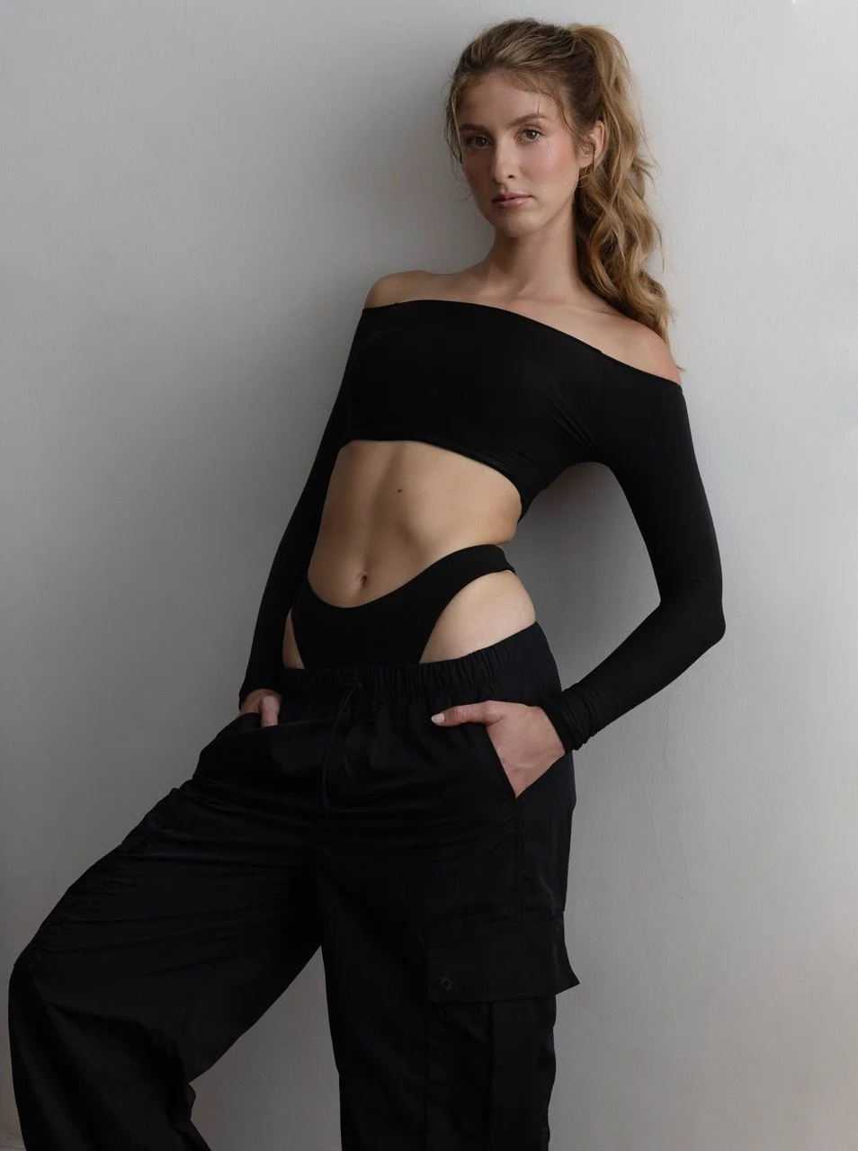 A young woman with red hair styled in loose waves, wearing a black off-the-shoulder crop top and black cargo pants, posing against a plain light gray wall.