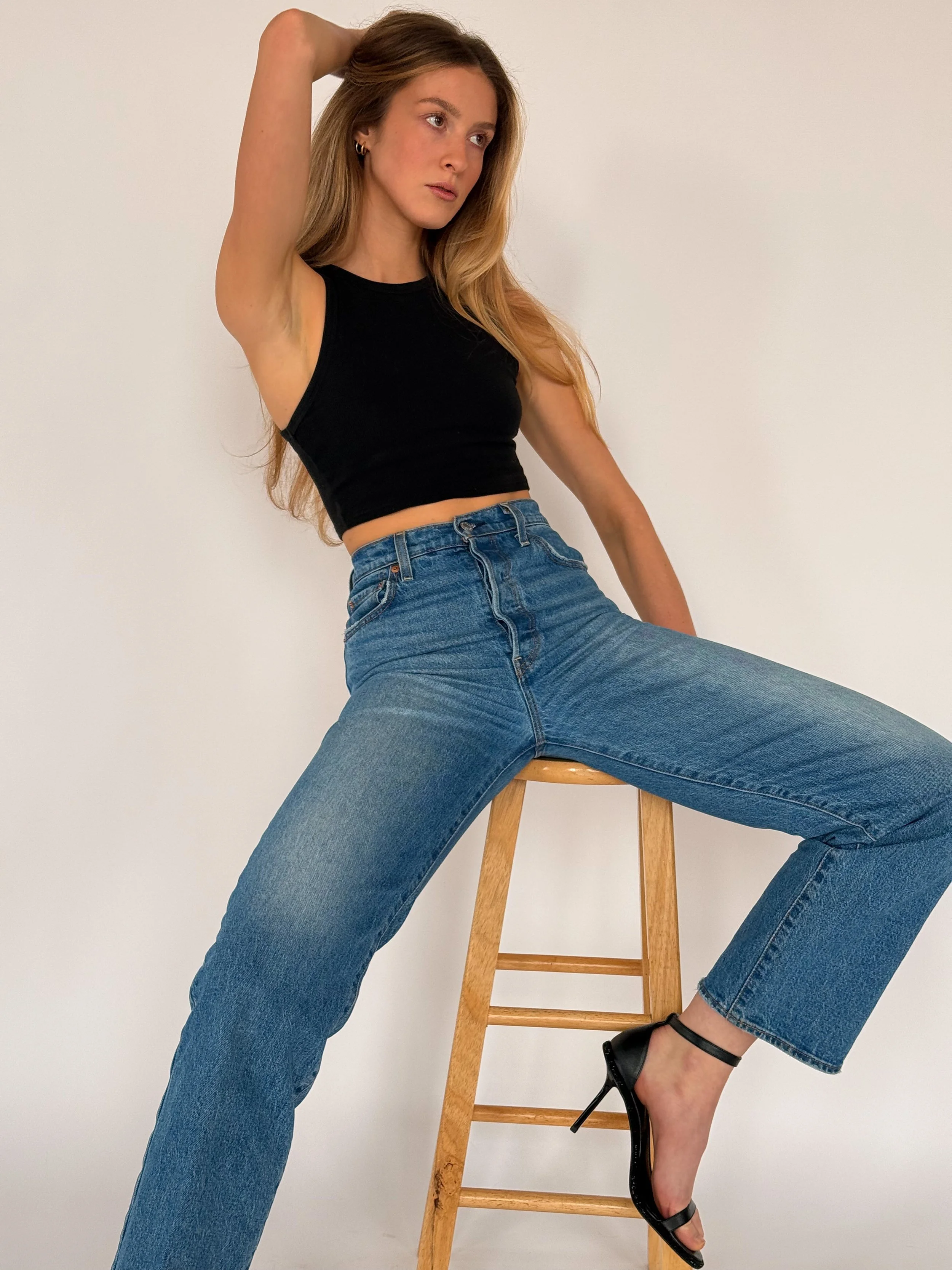 A woman with long blonde hair sitting on a wooden stool, wearing a black sleeveless crop top, high-waisted blue jeans, and black high-heeled sandals with ankle straps, posing against a plain white background.