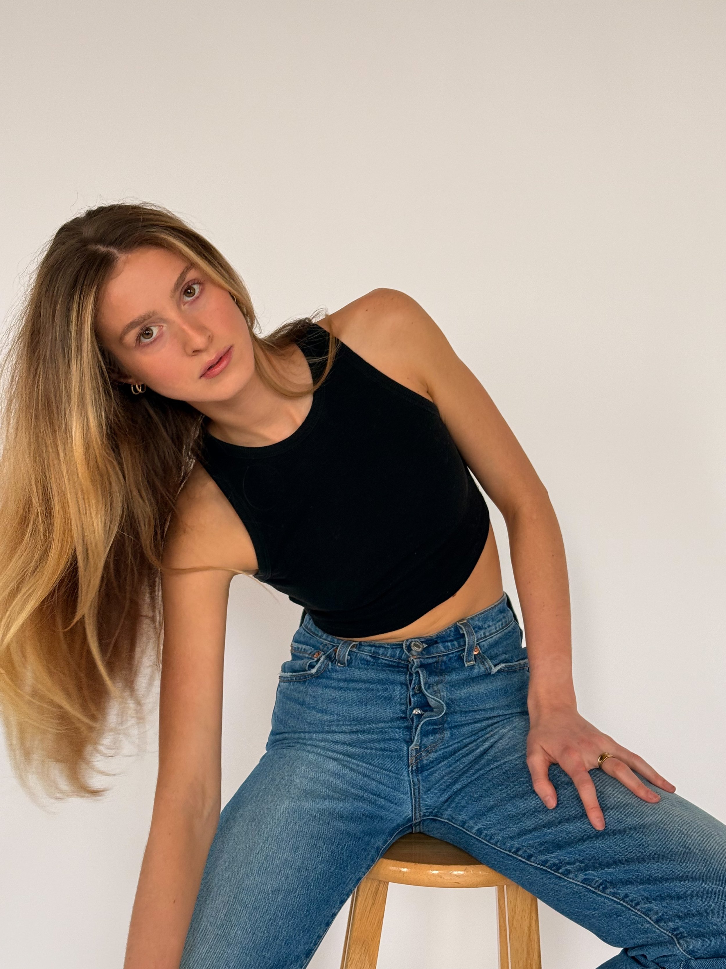 Young woman with long blonde hair wearing a black tank top and blue jeans sitting on a wooden stool against a white wall.