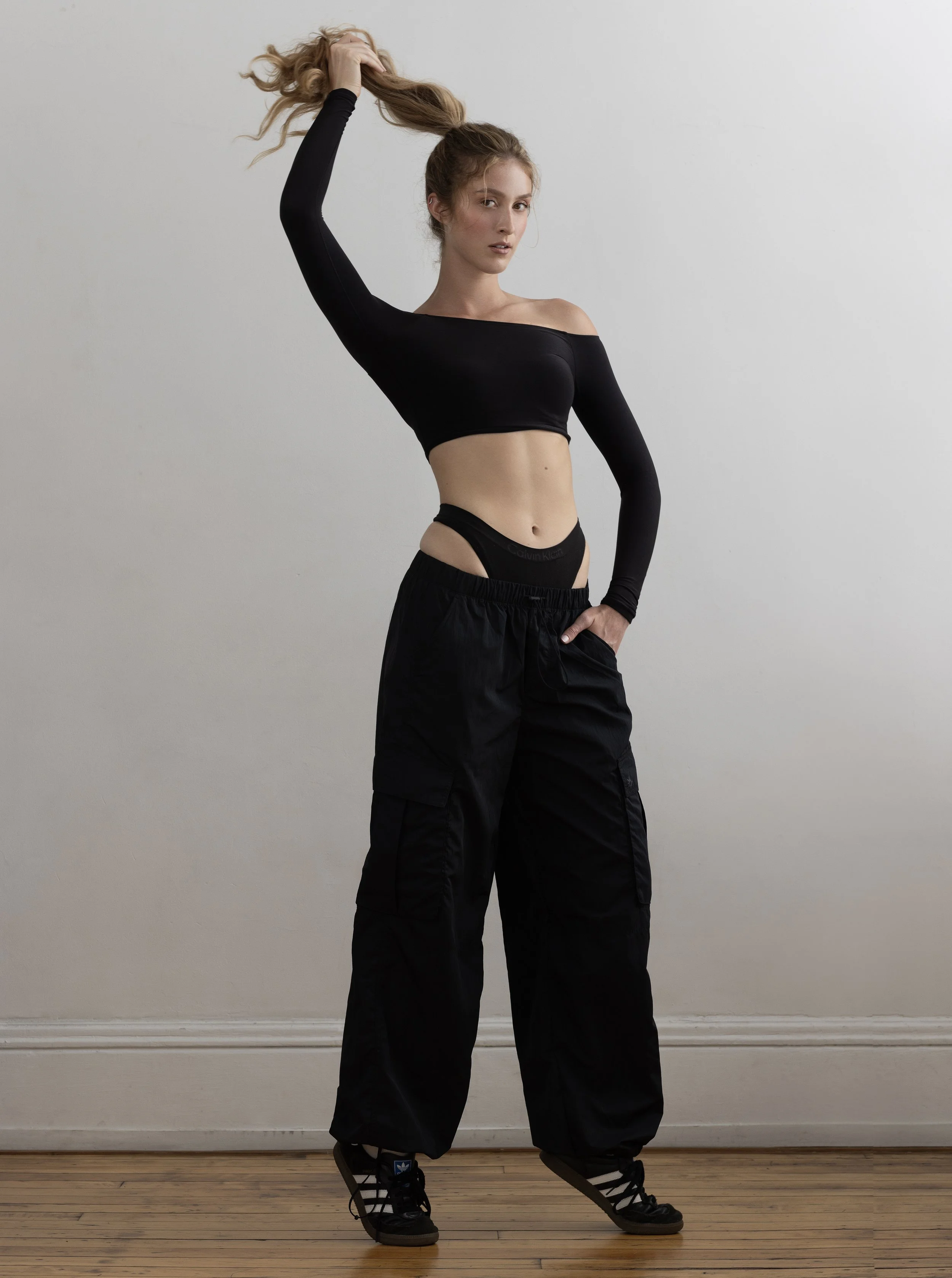 A young woman with long curly blonde hair, wearing a black long-sleeve crop top, black cargo pants, and black sneakers, standing against a plain light-colored wall, holding her hair in the air.