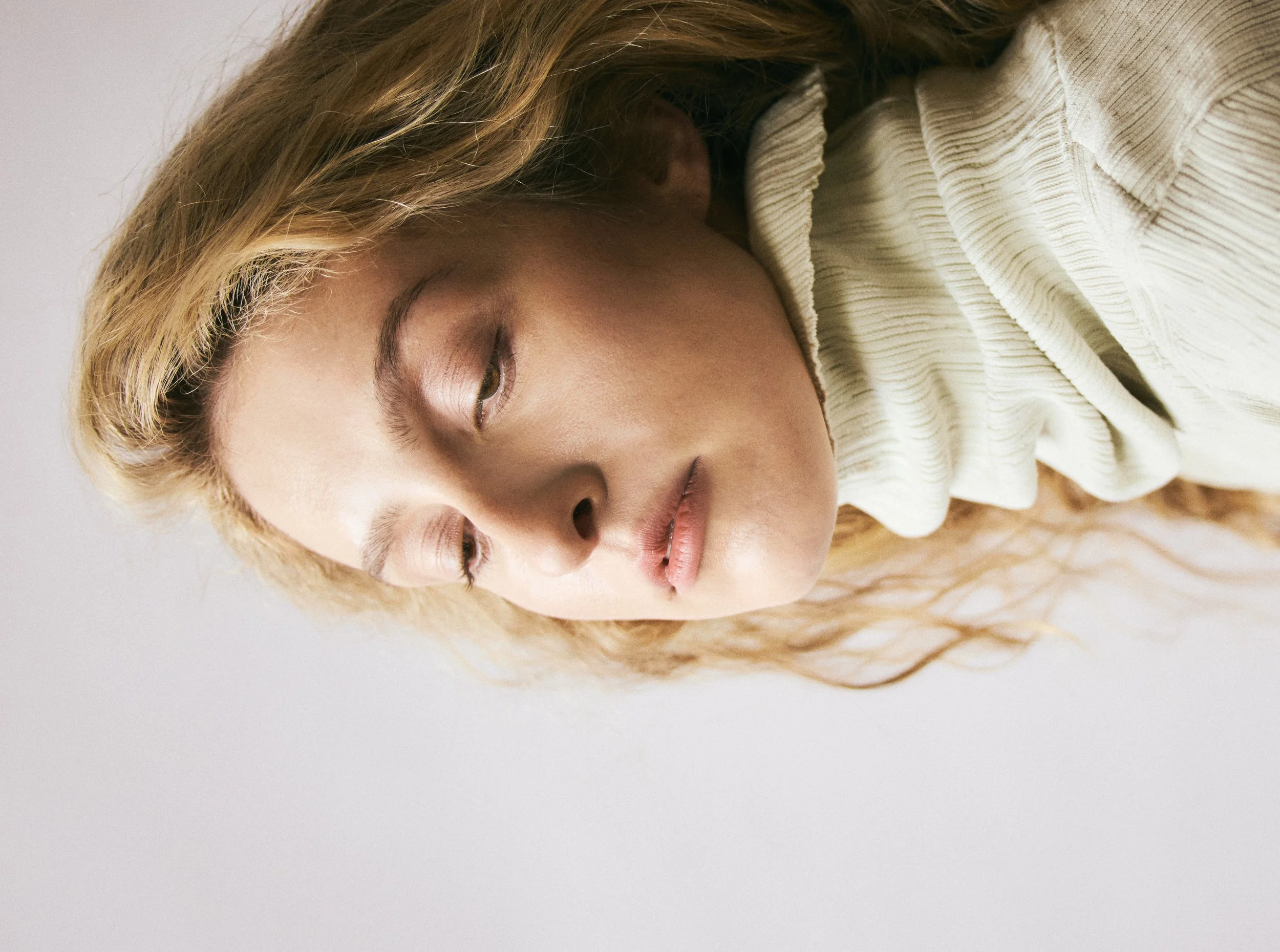 A close-up portrait of a young woman with wavy blonde hair, wearing a cream-colored textured top, with her head tilted and eyes closed against a plain background.