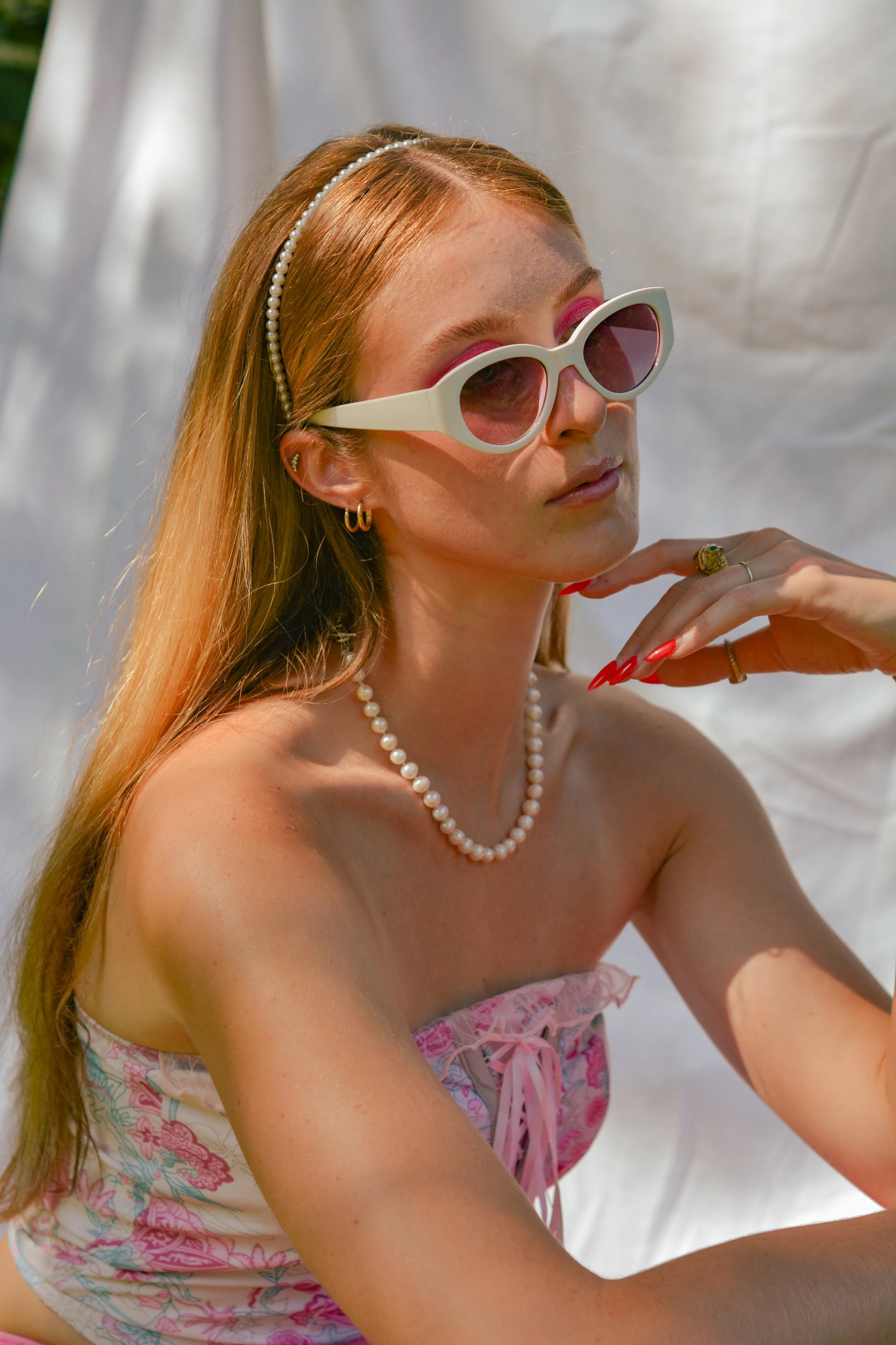 A young woman with long red hair wearing white sunglasses, pearl necklace, earrings, and a floral strapless top, posed outdoors in sunlight.