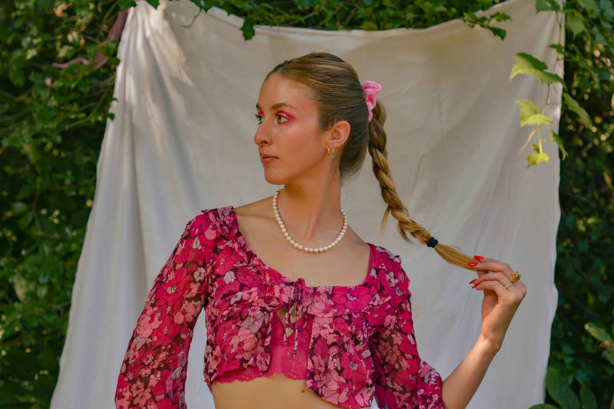 A woman with tan skin and long brown hair in a braid, holding the braid with one hand, wearing a pink patterned top, a pearl necklace, and gold jewelry, standing outdoors in front of a white fabric backdrop surrounded by greenery.