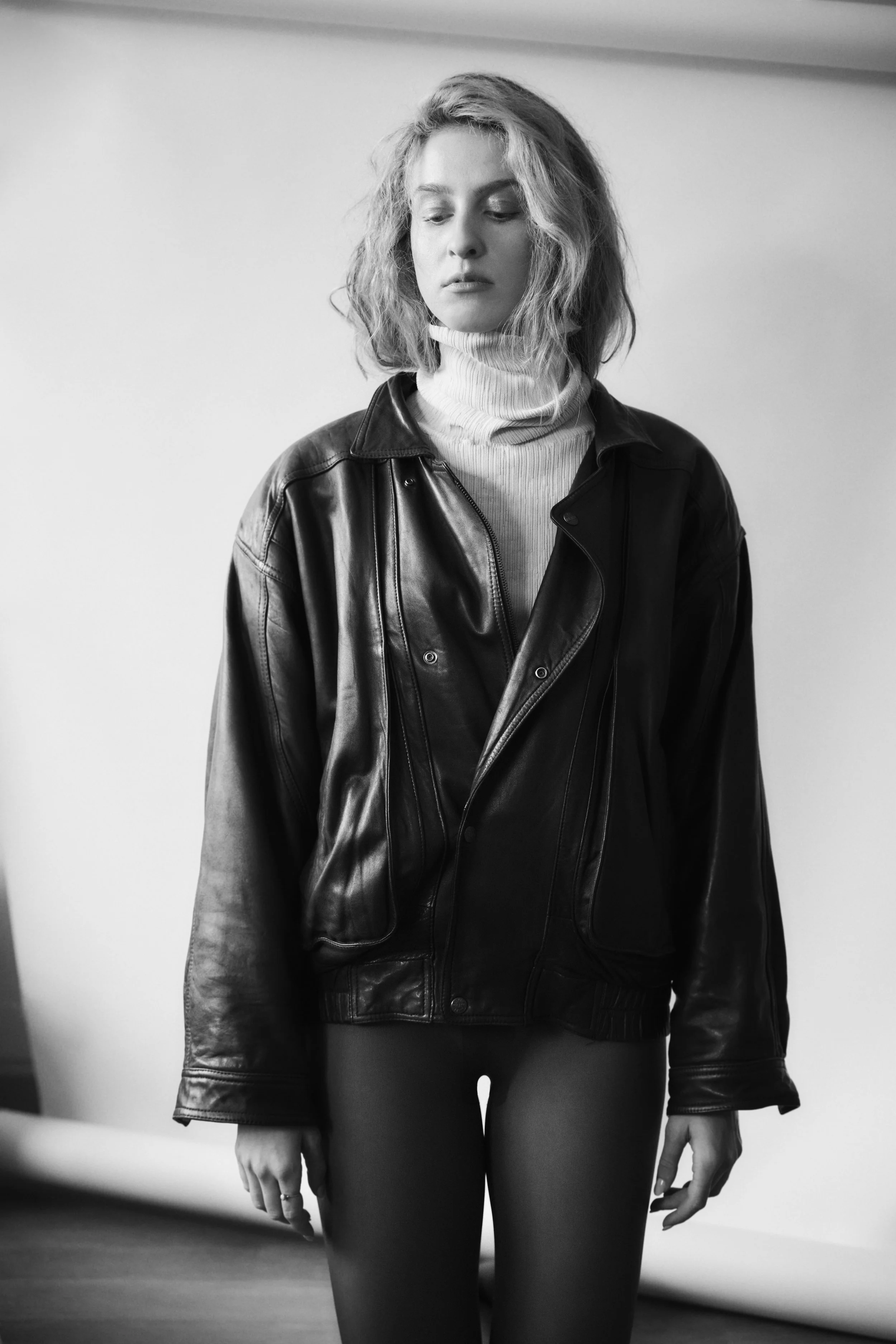 A young woman with wavy, shoulder-length hair wearing a leather jacket over a turtleneck shirt, standing indoors with a plain background.