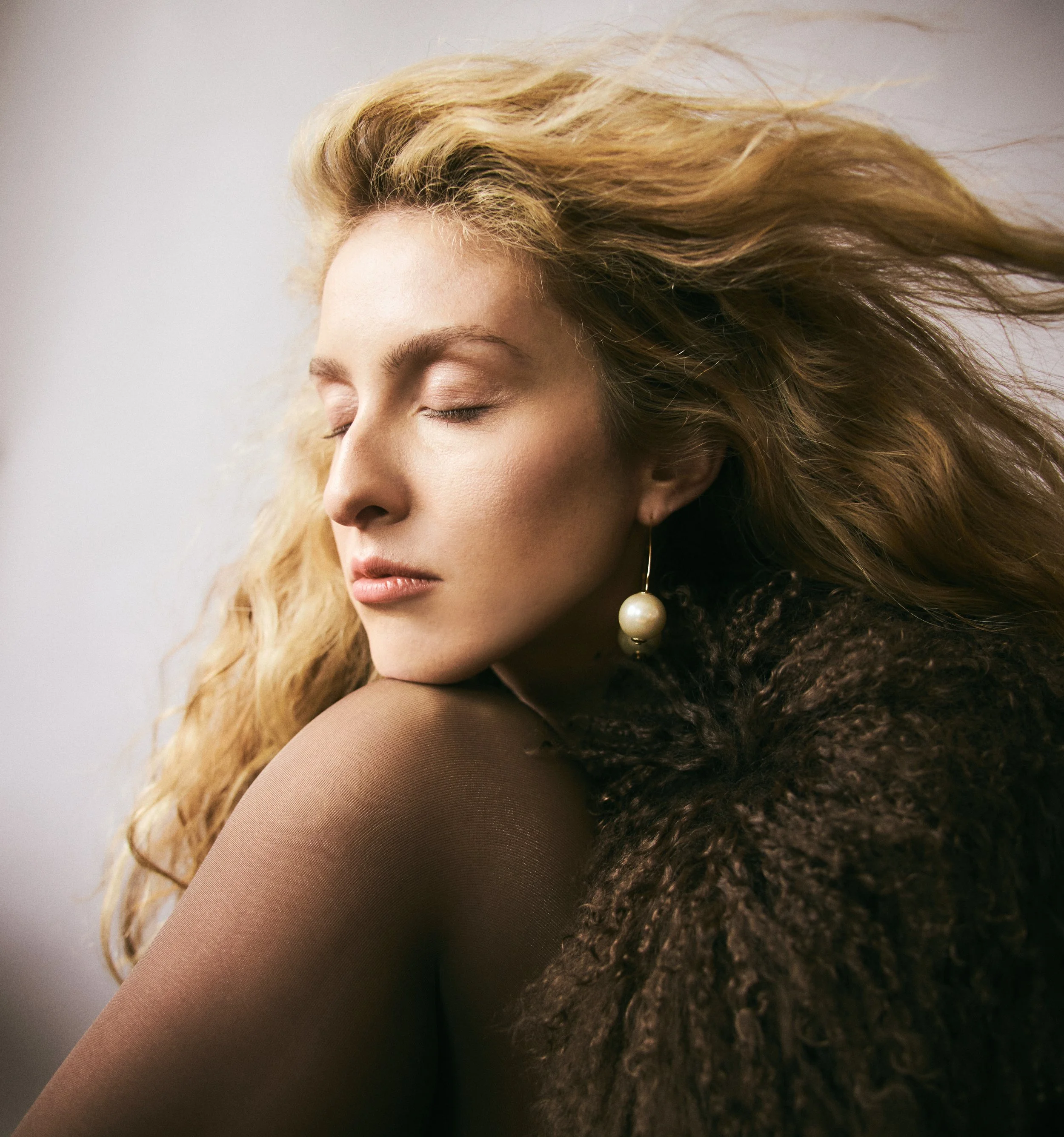 A woman with long, wavy blonde hair is in profile with her eyes closed, wearing a large pearl earring and a cozy dark brown fur coat.