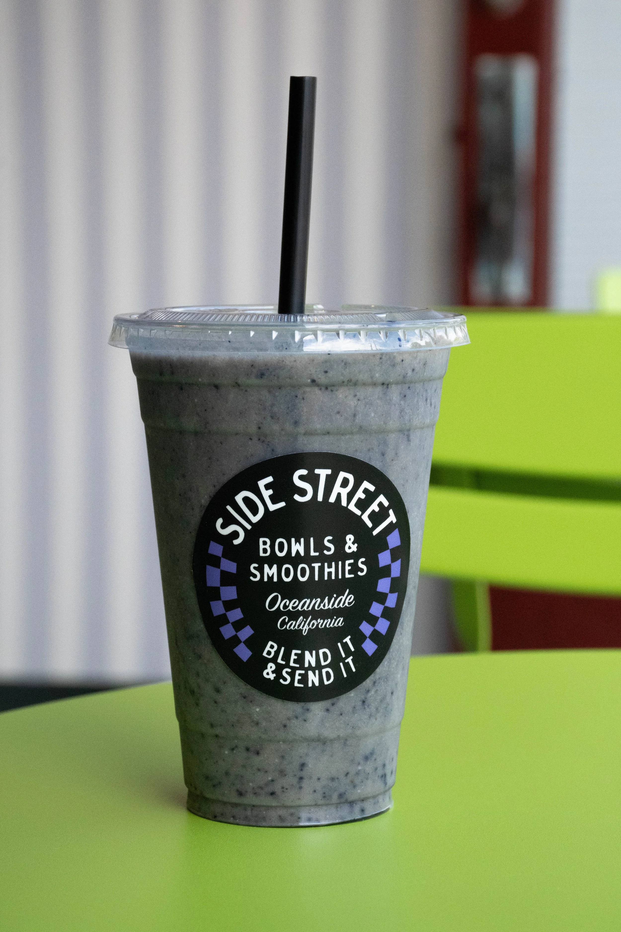 A smoothie in a plastic cup with a black straw, placed on a lime green table, with a black and white label reading 'Side Street Bowls & Smoothies Oceanside California.'  @laughingcoyote24