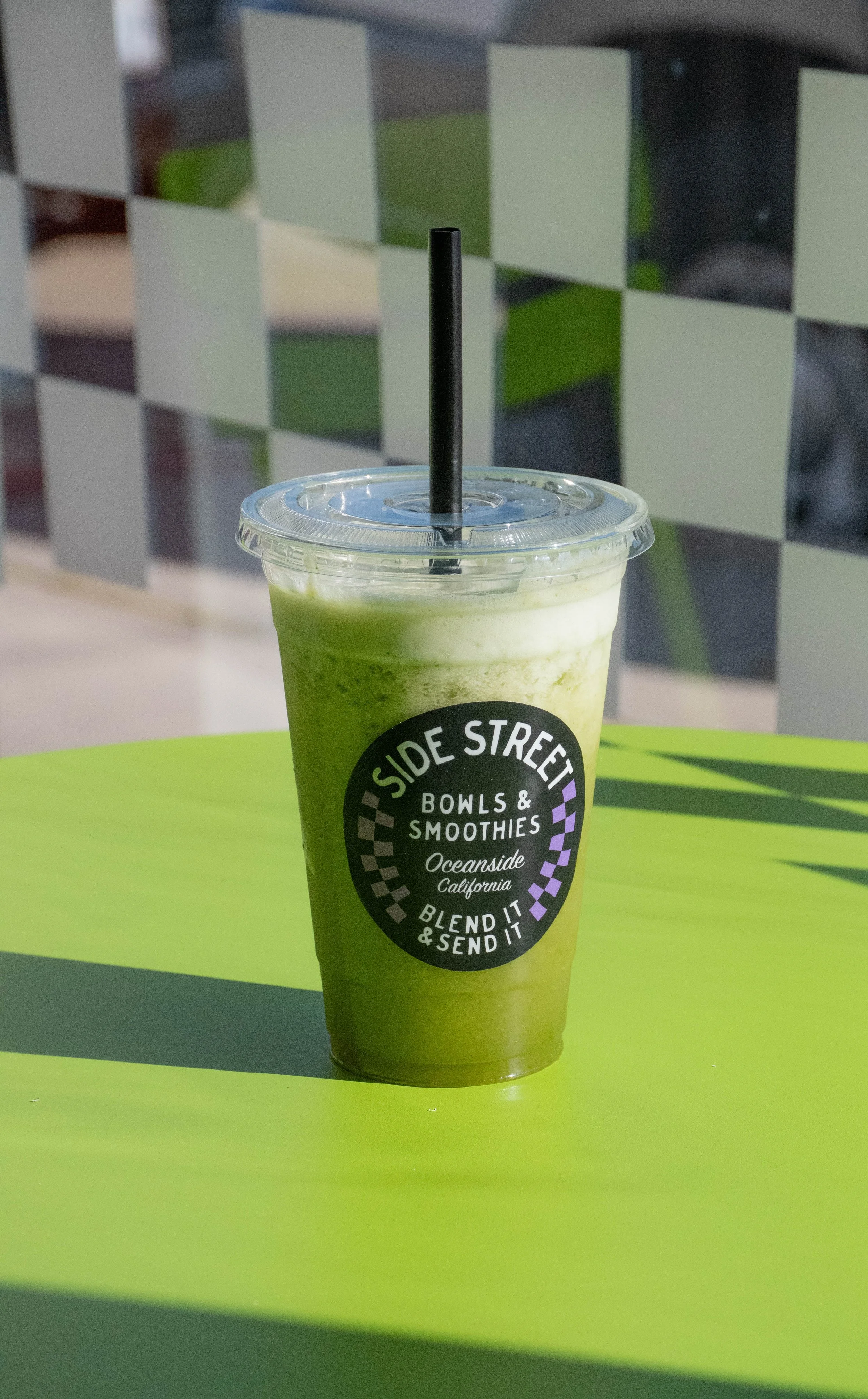 A green smoothie in a clear plastic cup with a black straw, placed on a bright green table. The cup has a logo for Side Street Bowls & Smoothies, located in Oceanside, California.  @laughingcoyote24