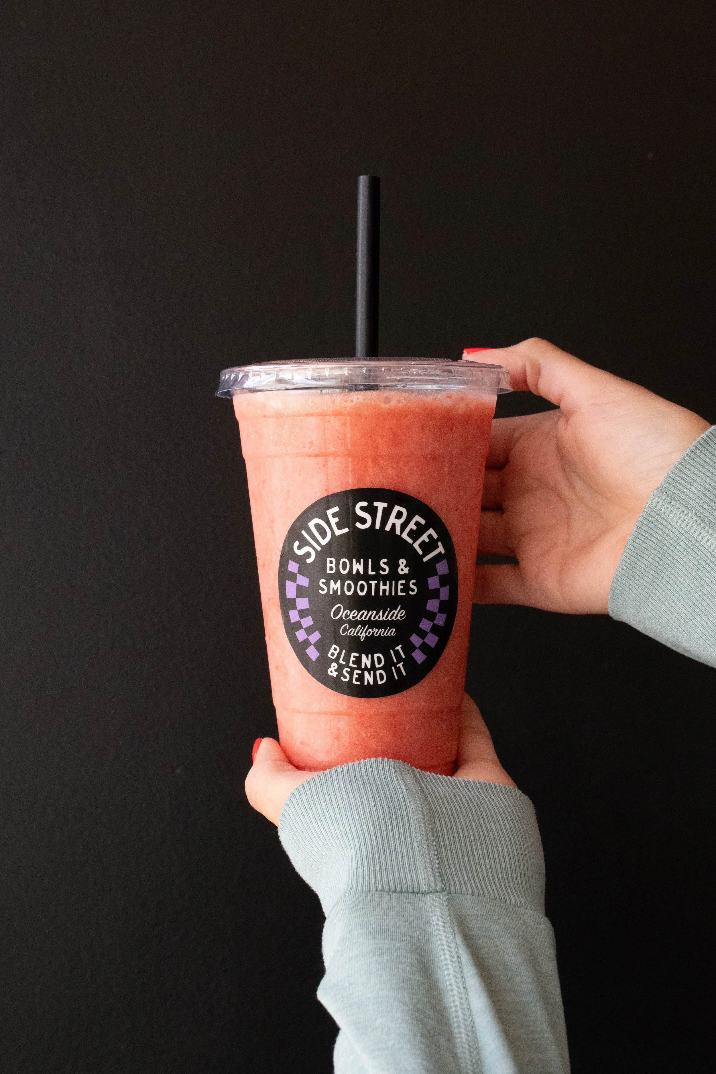 A hand holding a large cup of pink smoothie from Side Street Bowls & Smoothies in Oceanside, California, with a black straw, against a black background.  @laughingcoyote24