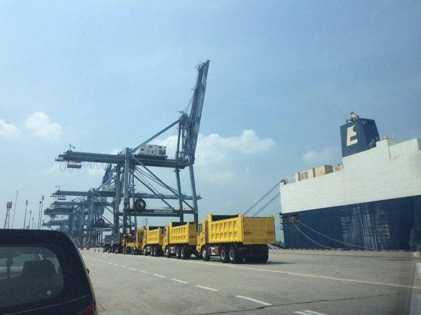 Cargo trucks parked beside container cranes and a large ship at a busy seaport.