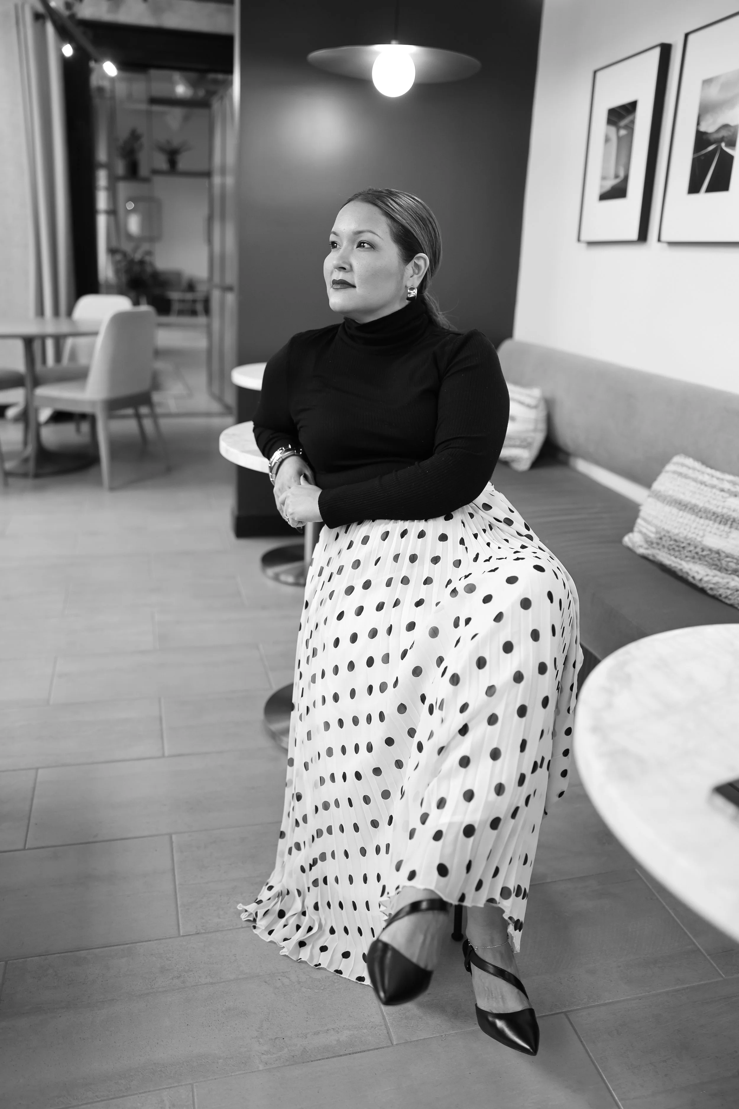 A woman sitting on a bench in a modern indoor space, wearing a black turtleneck, polka dot skirt, and heels, with framed pictures on the wall behind her.