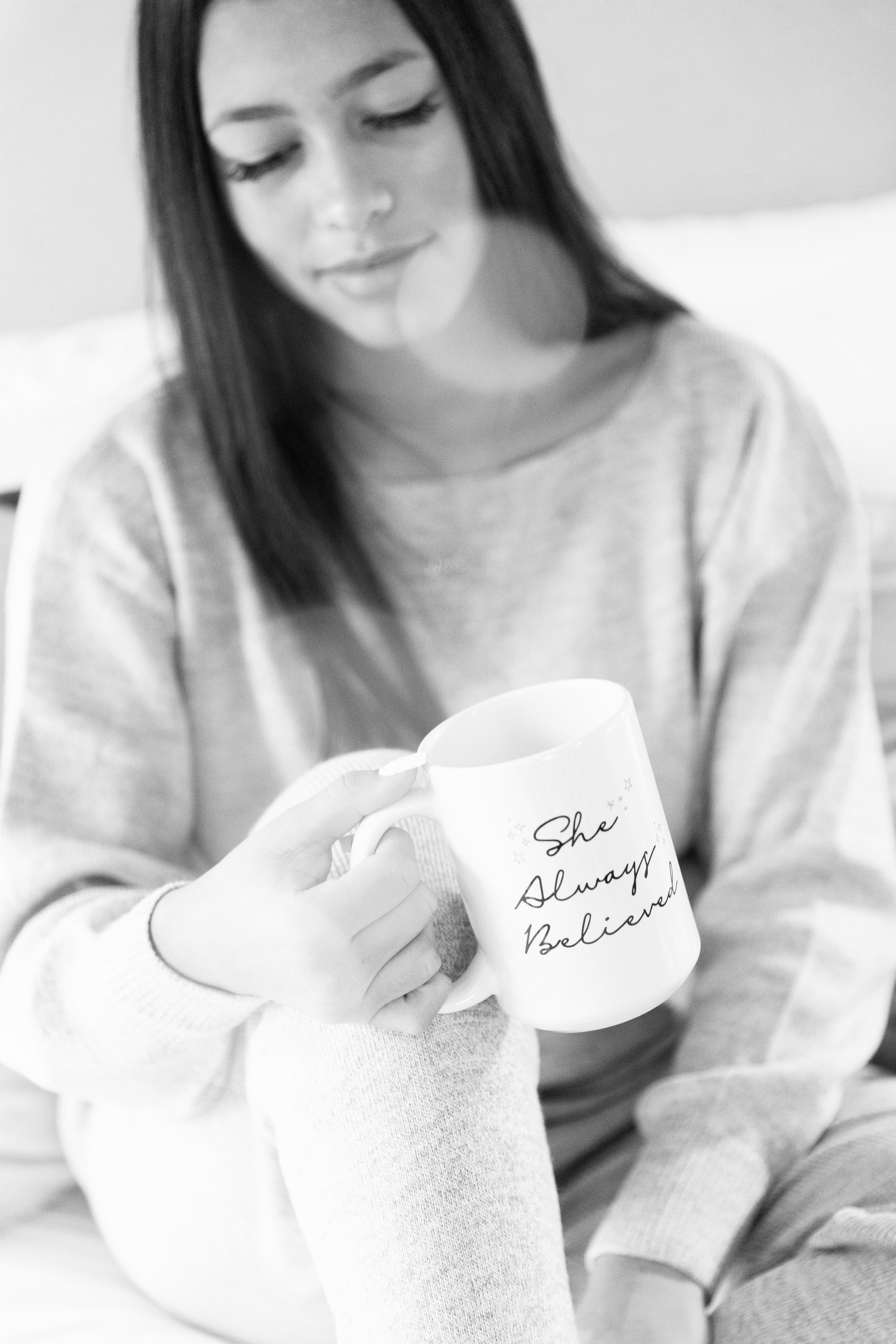 A young woman with straight dark hair and a relaxed expression, holding a white mug with the words "She Always Believed" written on it in cursive, in a cozy indoor setting.
