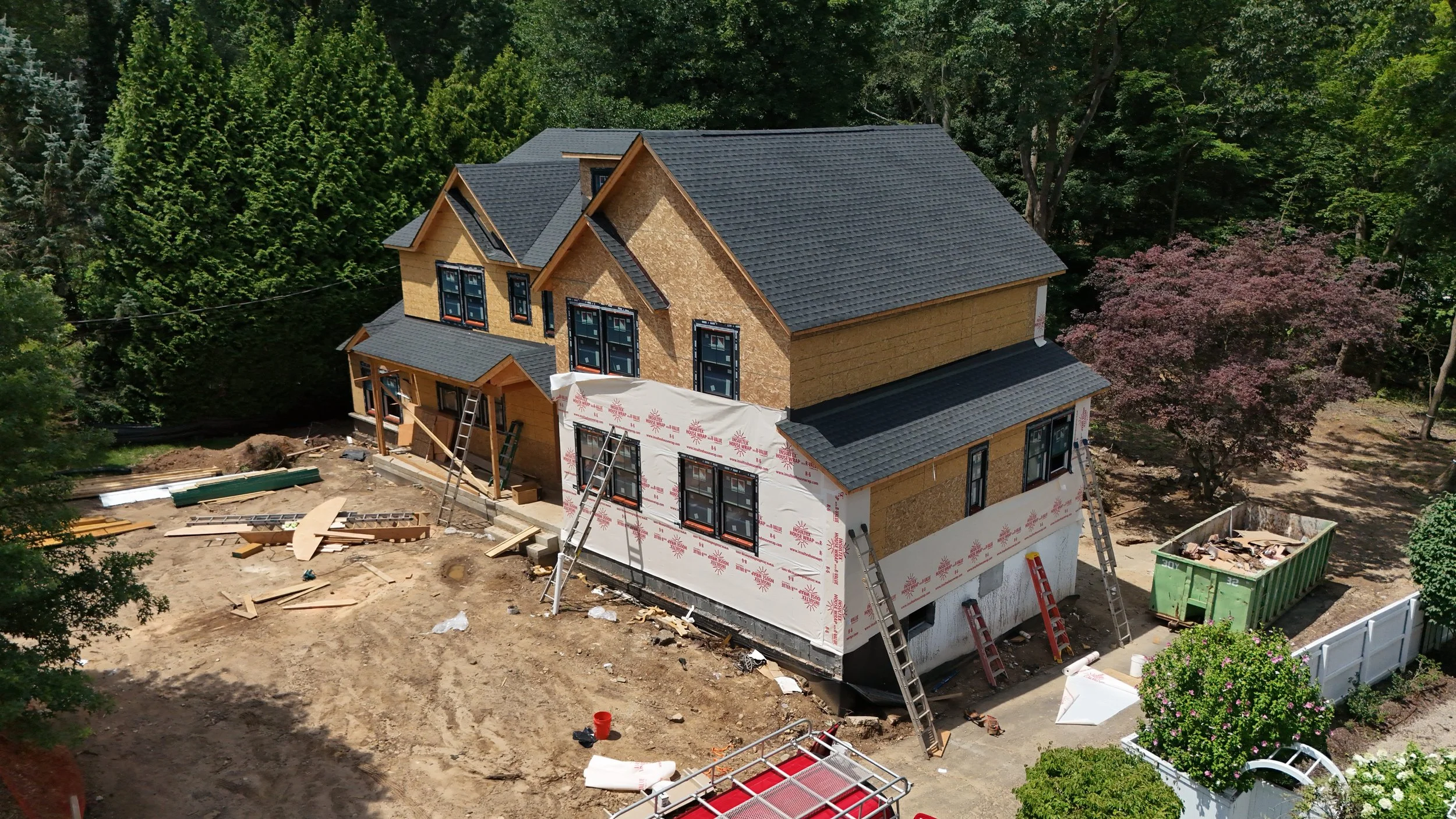 A house under construction with multiple floors and a dark gray roof, surrounded by trees. Construction materials and ladders are visible on the site, with some walls partially covered in white weatherproofing material.