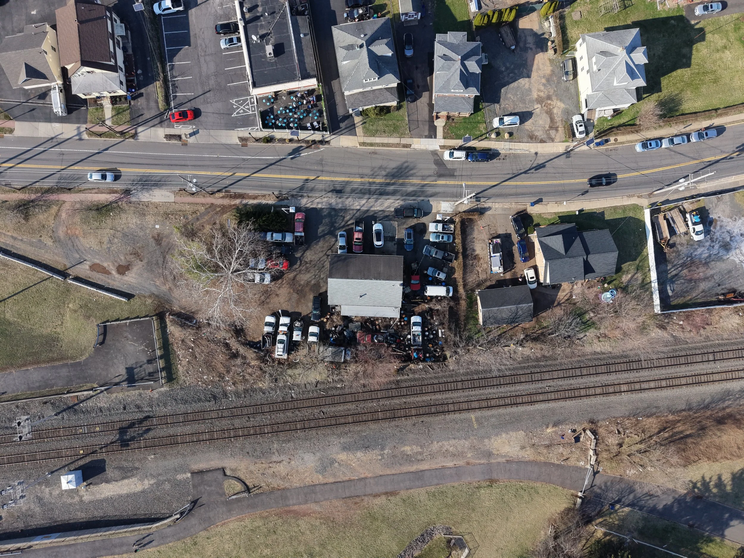 Aerial view of a neighborhood with houses, cars parked along the streets, and a railway line at the bottom of the image. There is a mix of residential buildings and open spaces.