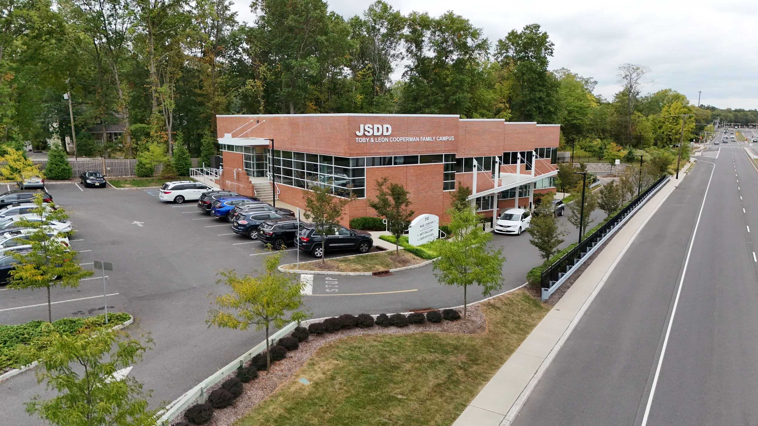 Aerial view of a brick building labeled "JSDD Toby & Leon Cooperman Family Campus" with a parking lot filled with cars surrounded by trees, next to a multi-lane road with minimal traffic.