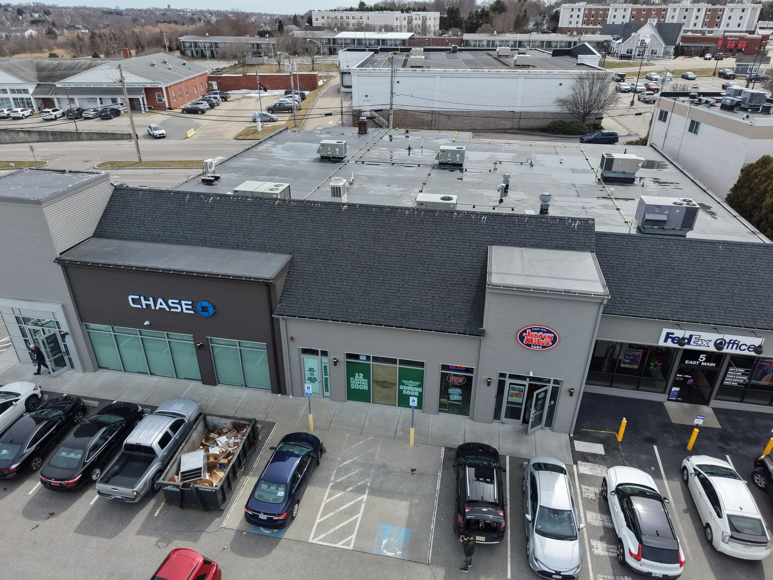 A parking lot in front of a commercial building featuring a Chase bank branch, Jersey Mikes, and FedEx Office store, with parked cars and a dumpster with trash.