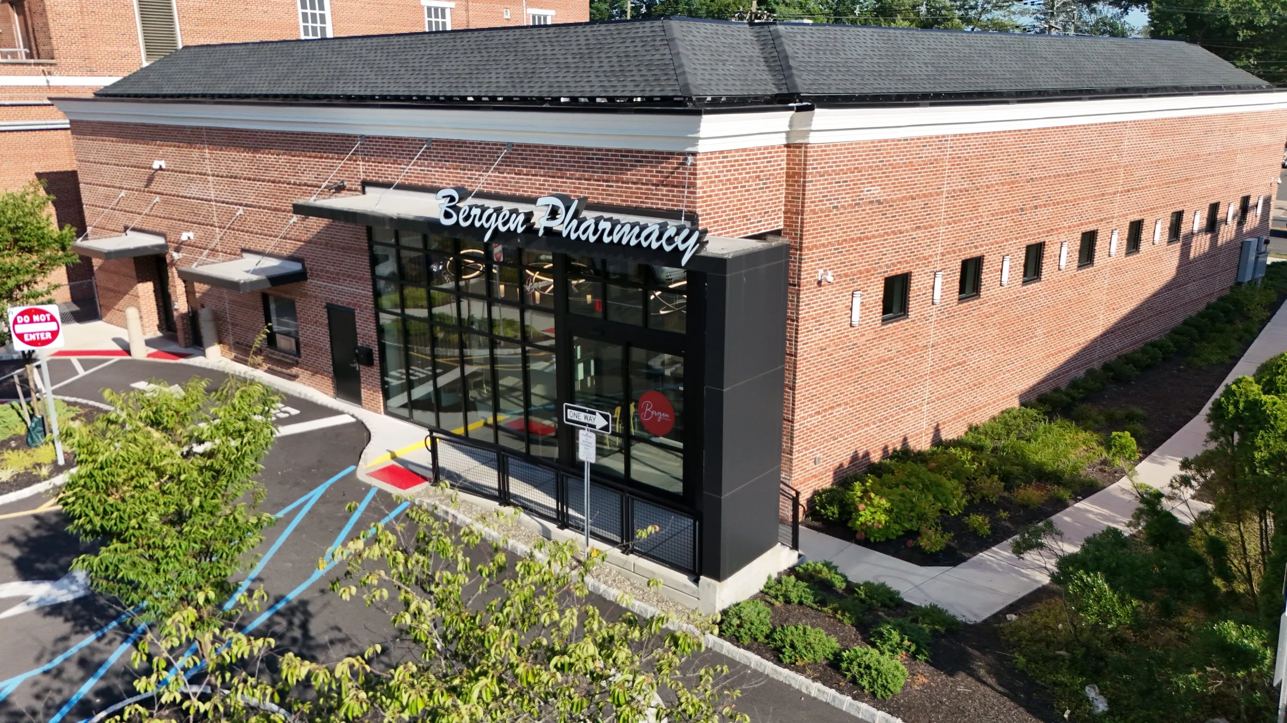 Exterior of Bergen Pharmacy with a brick building, black entrance, and parking lot with blue and white lines, low shrubbery, and trees.