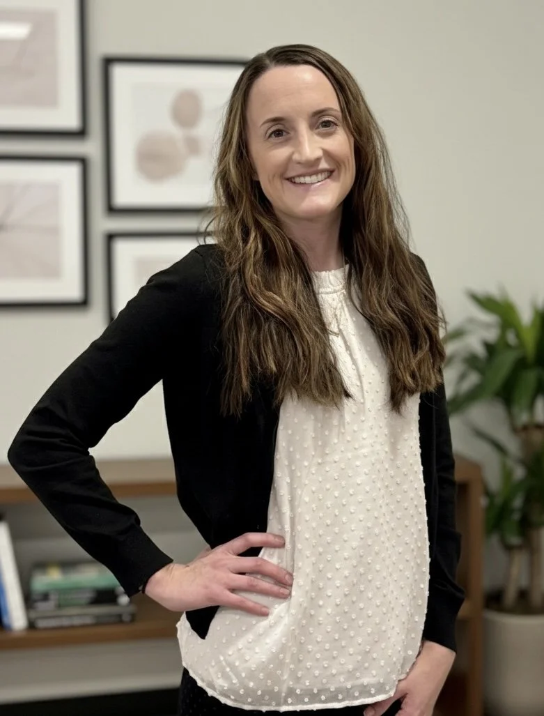 A woman with long, wavy brown hair and a bright smile stands in an office, wearing a black blazer over a white polka dot blouse. She has one hand on her hip and is posing in front of framed artwork and a potted plant.
Therapist in a pediatric clinic.