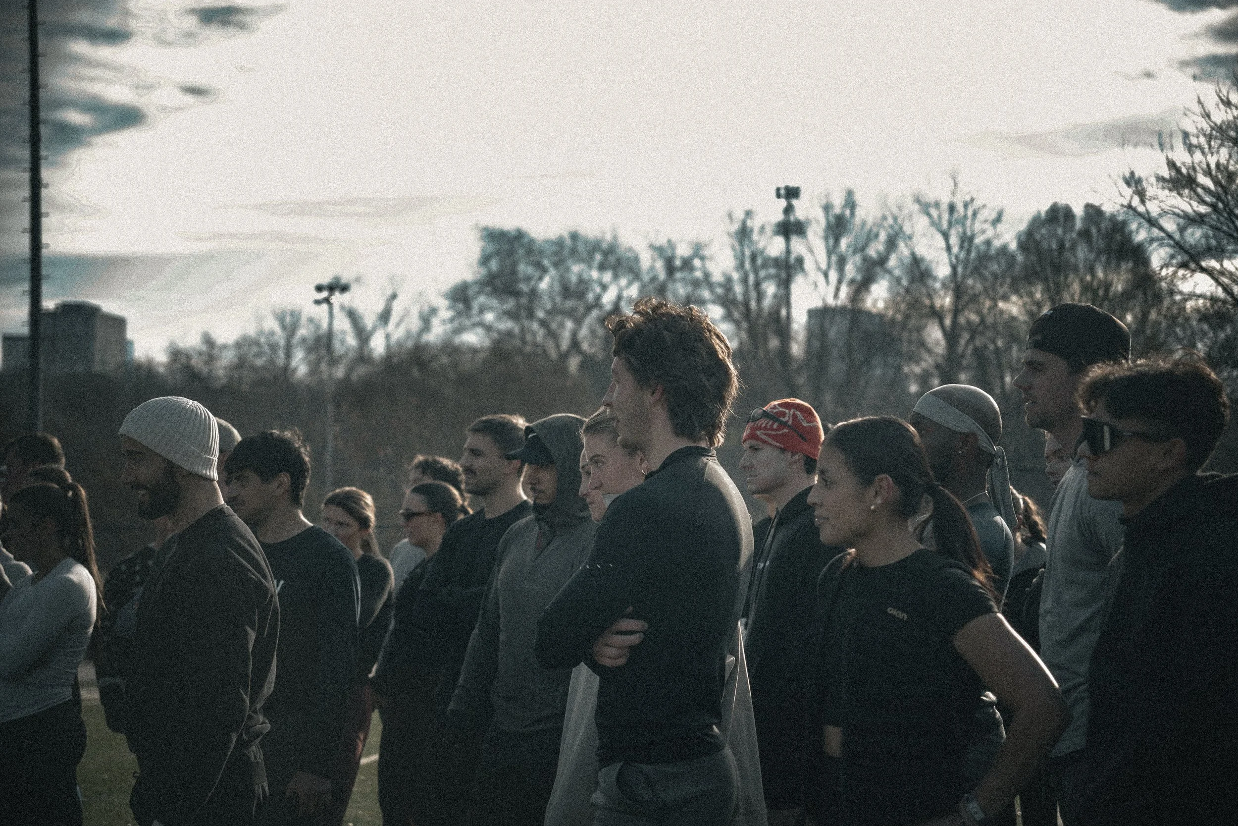 A group of diverse people standing outdoors during daytime, some wearing athletic clothing and accessories, in an open area with trees and power lines in the background.