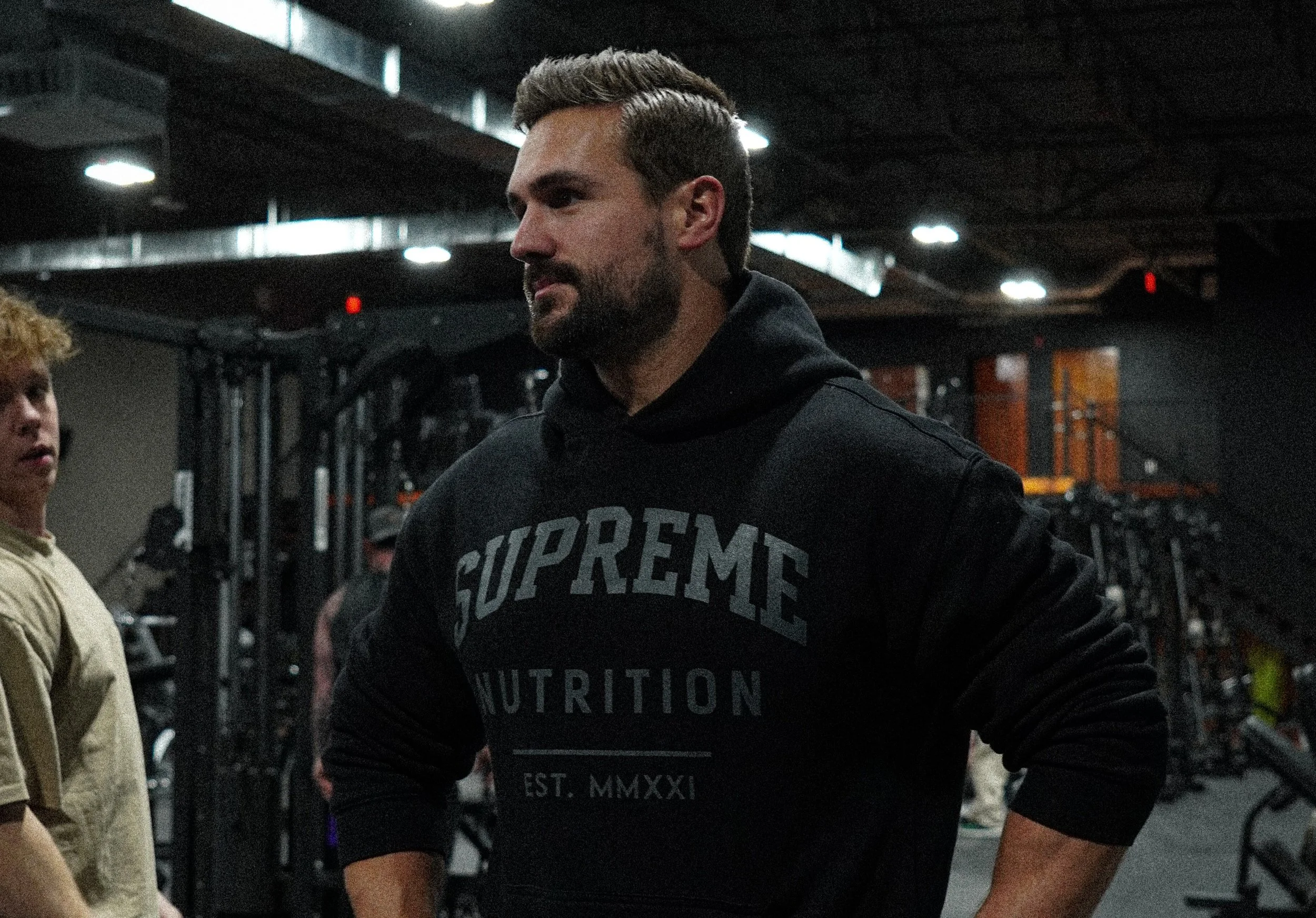 A man with a beard and short hair, wearing a black hoodie with 'SUPREME NUTRITION' printed on it, standing in a gym with exercise equipment in the background.