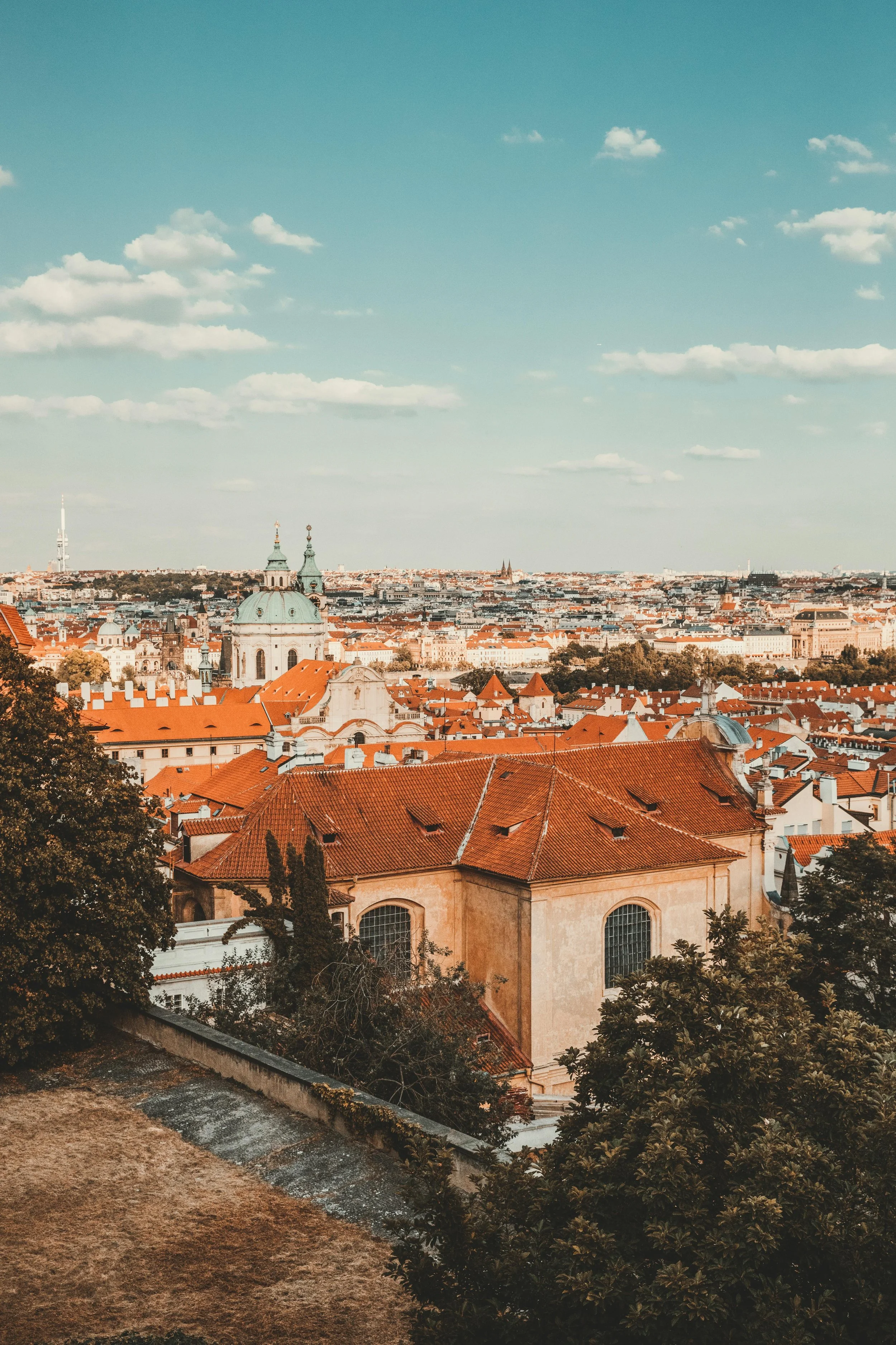 A cityscape of Prague with orange rooftops, church domes, and a distant view of the city under a blue sky with scattered clouds.
