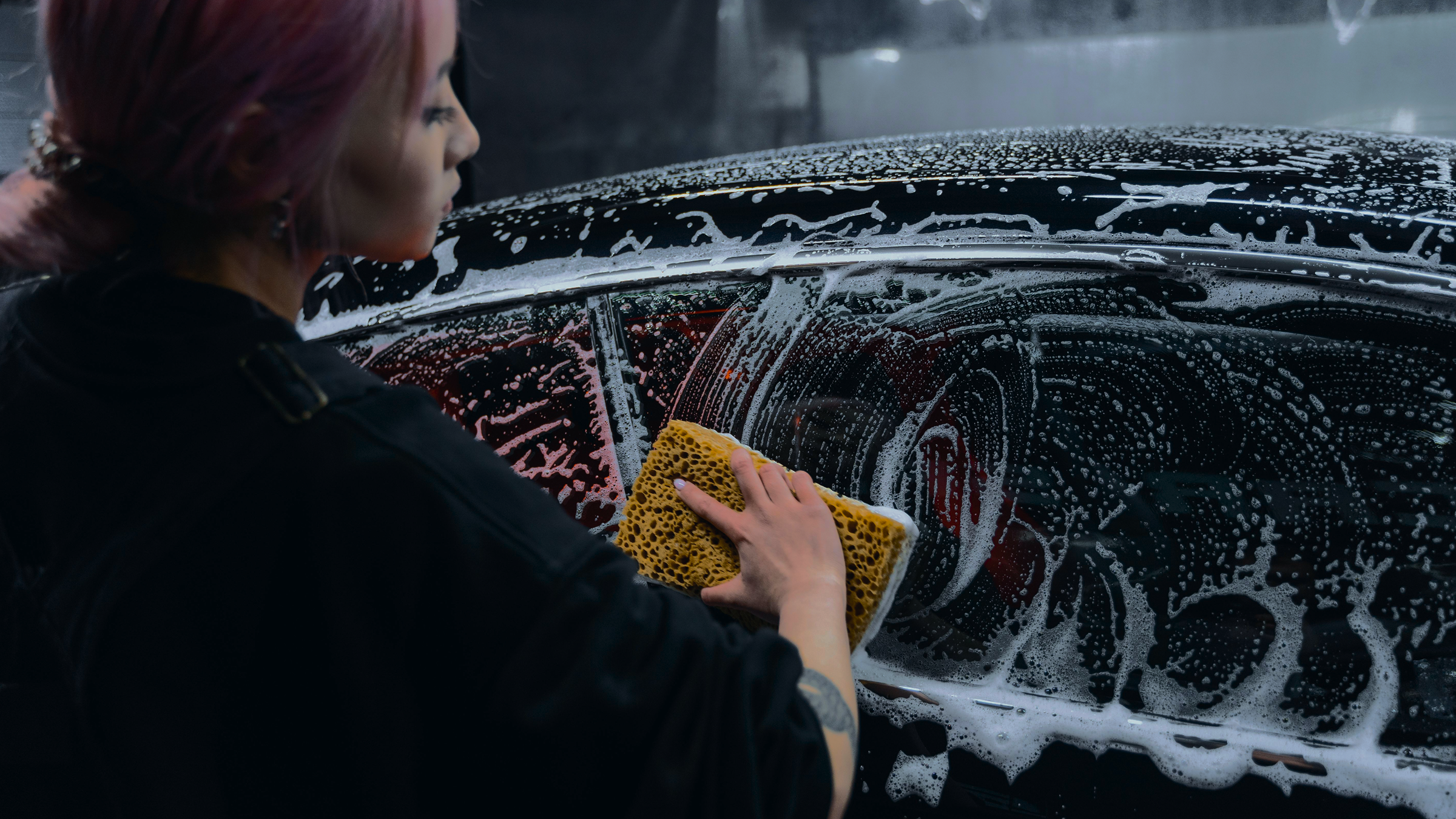 A woman with purple hair wearing a black jacket washing a black car with a yellow sponge.