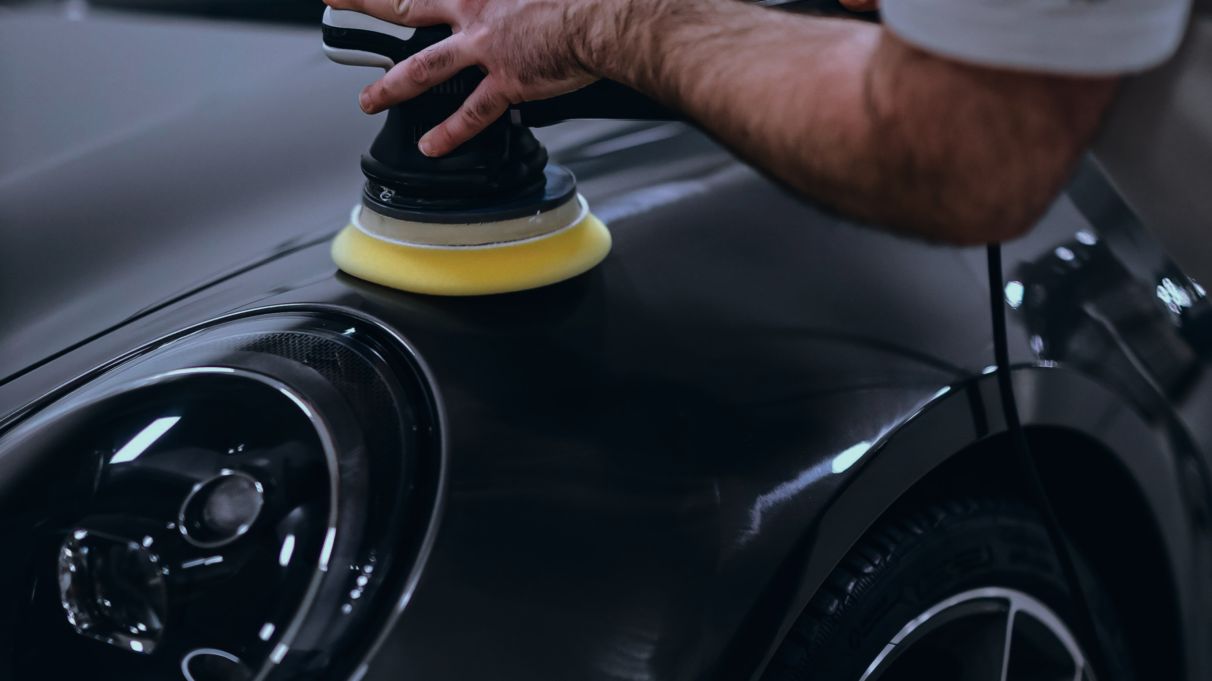 Person polishing a black car with a yellow foam pad and a power buffer in a garage or detailing shop.