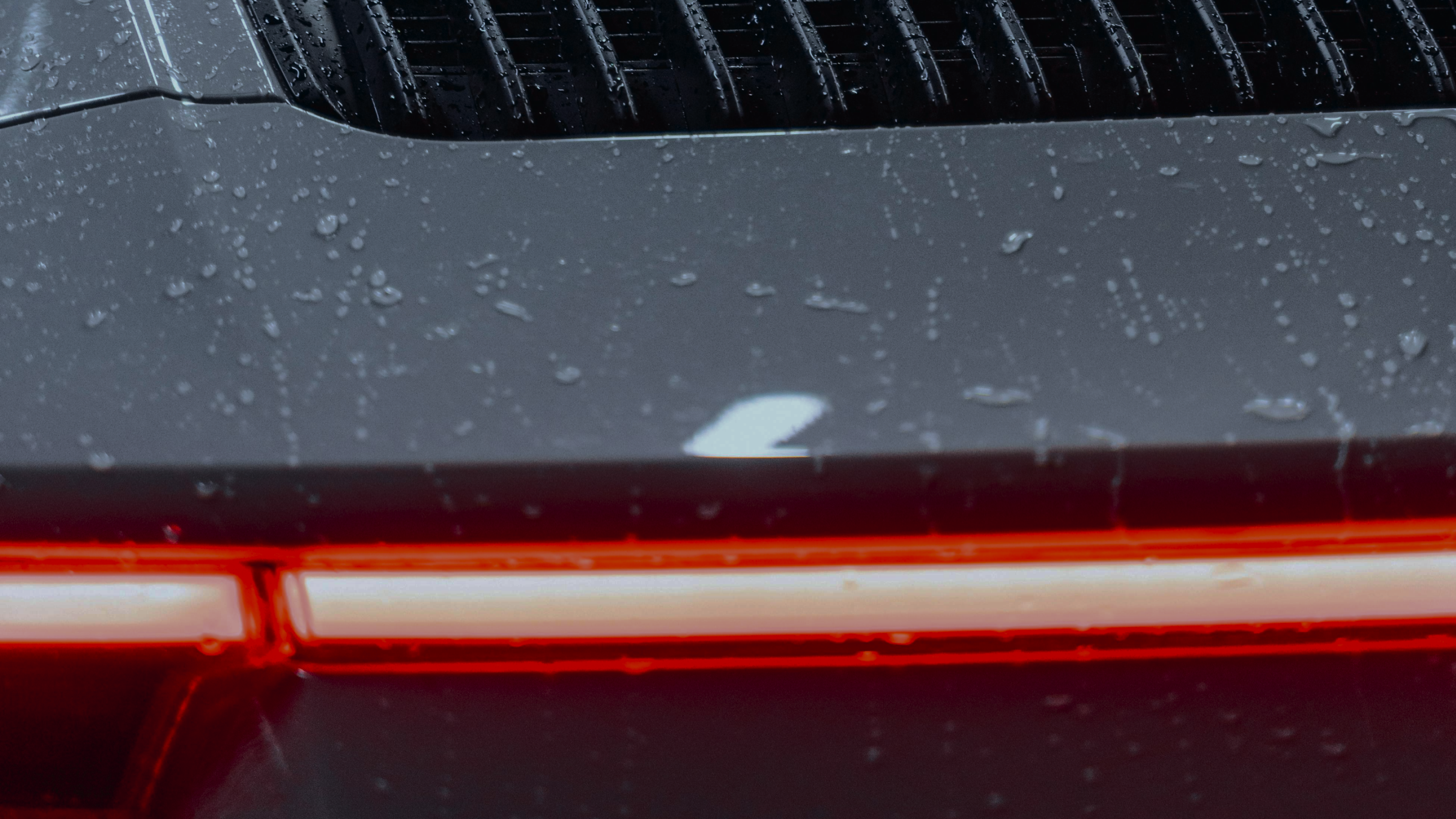 Close-up of the rear of a dark-colored car, focusing on the taillight and wet surface with raindrops.