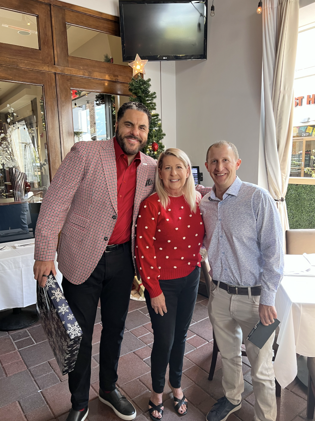 Three people, two men and one woman, standing together in a restaurant with a Christmas tree in the background. The man on the left is wearing a red shirt with a patterned jacket, the woman in the middle is in a red sweater with white polka dots, and the man on the right is dressed in a light-colored shirt. They are smiling and posing for the photo.