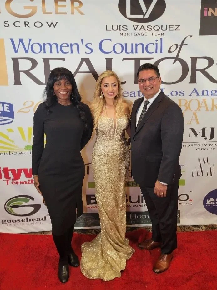 Three formally dressed people standing on a red carpet in front of a backdrop with various sponsor logos, including Women's Council of REALTORS, Luis Vasquez, and others. The woman in the middle is wearing a gold gown, the woman on the left is in a black dress, and the man on the right is in a dark suit with glasses.