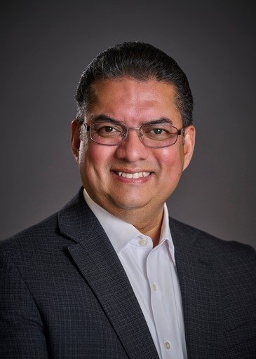 Professional headshot of a man with dark hair, glasses, wearing a dark suit jacket and white shirt, smiling against a dark background.