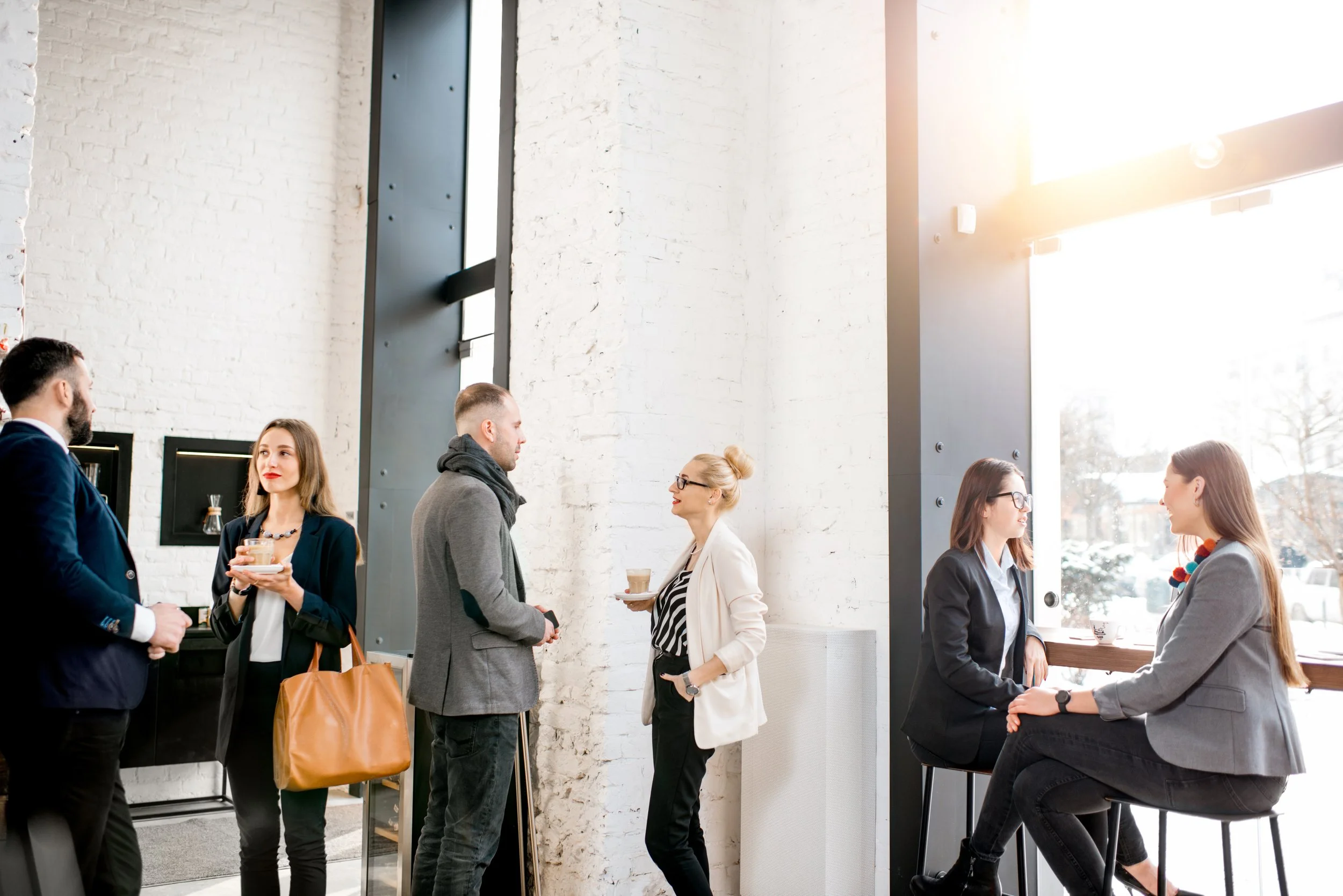 Group of people talking and enjoying coffee inside a bright, modern cafe with white brick walls and large windows.