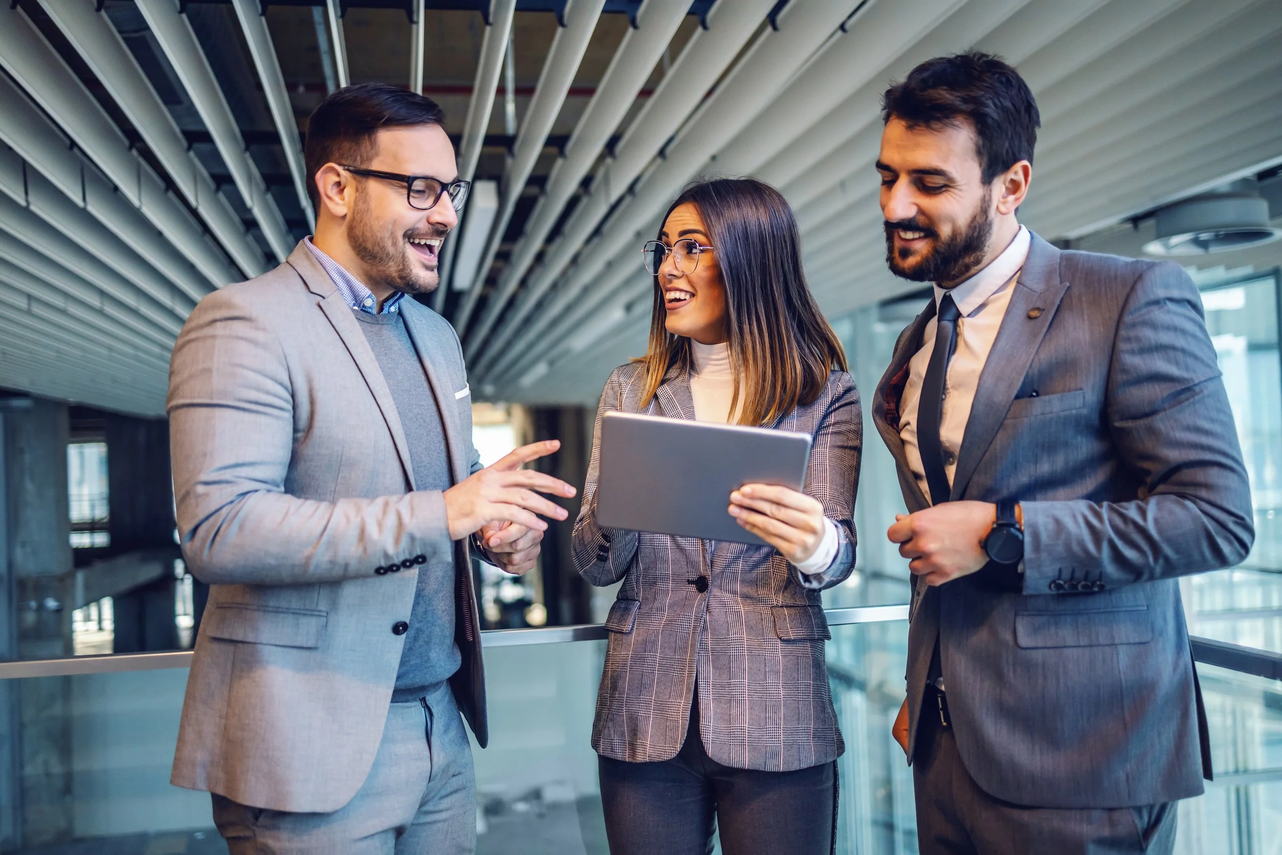 Three business professionals, two men and one woman, standing together in a modern office building, smiling and looking at a tablet held by the woman.
