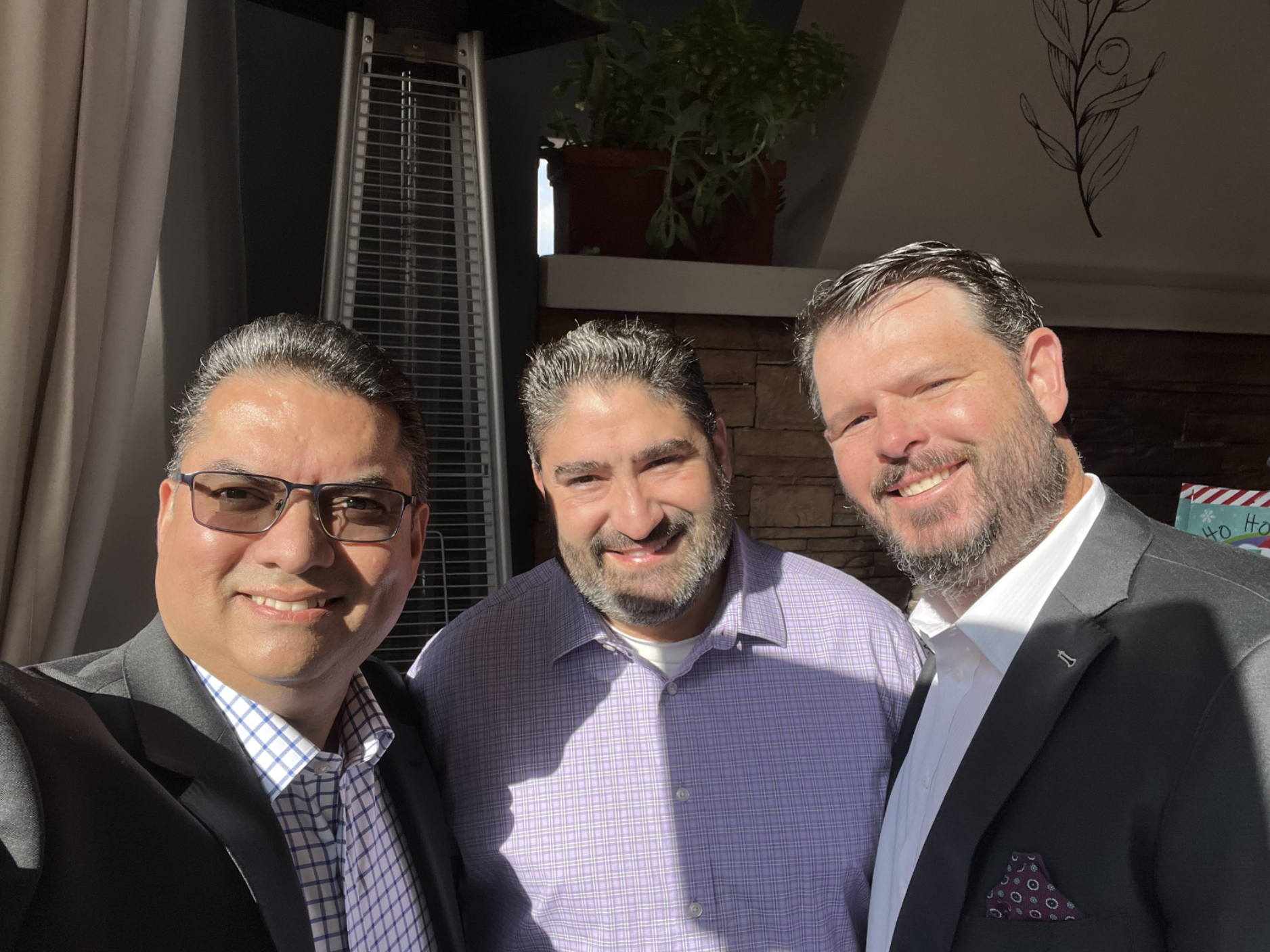 Three men smiling and posing for a photo indoors, dressed in business attire, with a plant and wall decor in the background.