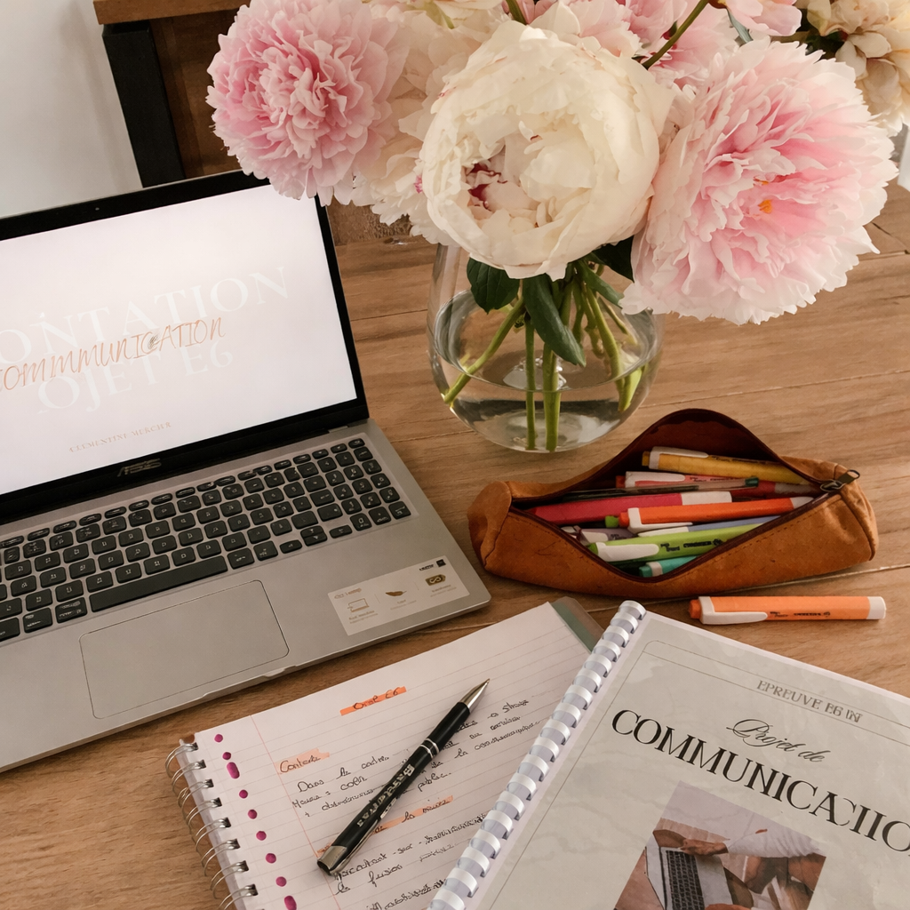 A workspace with a laptop displaying the word 'Communication', a vase of pink and white peonies, a leather pencil case with colored pens, and a notebook with handwritten notes. A printed booklet titled 'Prova de Comunicação' is also on the table.