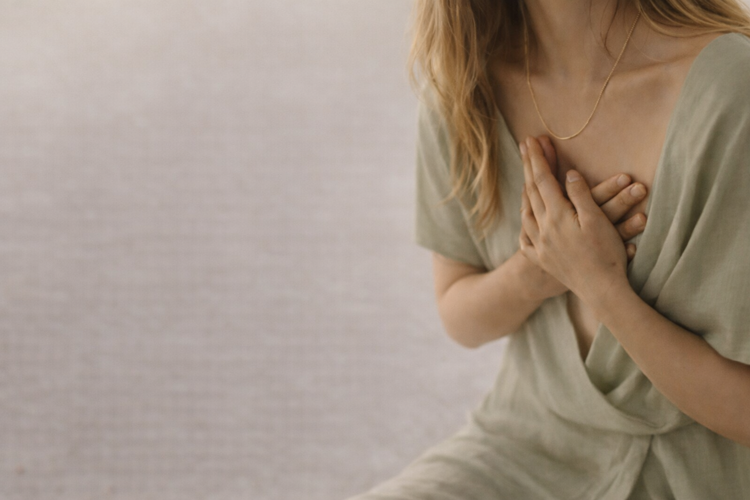 A woman with long blonde hair, wearing a gold necklace and a beige top, holds her hands over her chest.