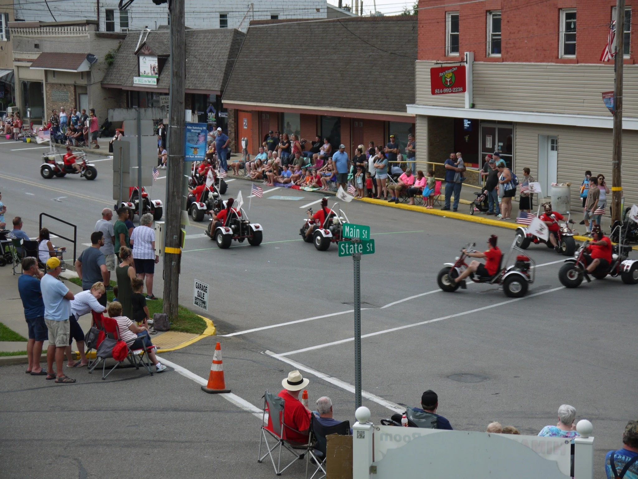 Parade on Main Street with decorated trikes, spectators seated and standing on sidewalk, and people watching from chairs and benches.