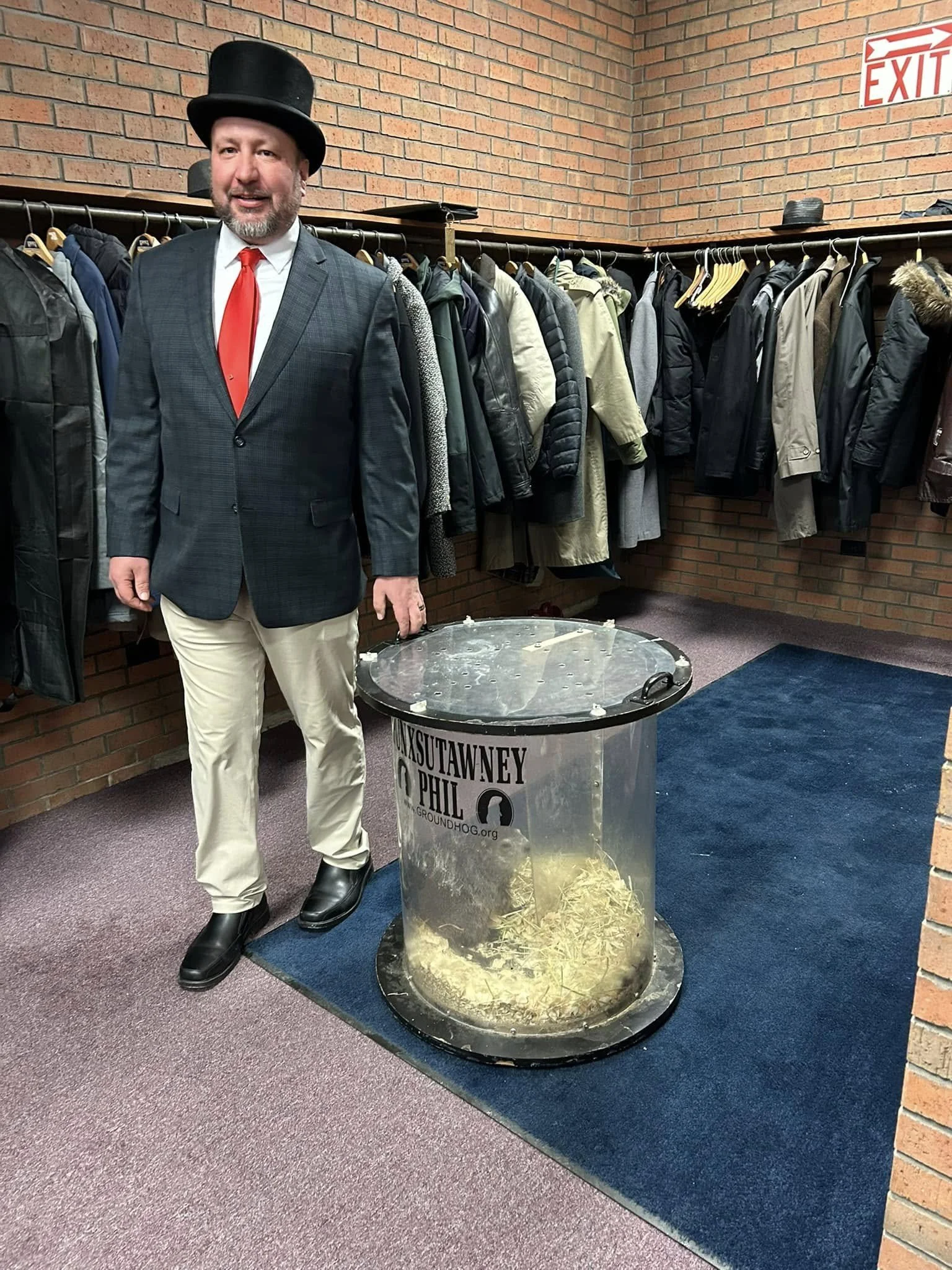 A man dressed in a suit, red tie, and top hat standing indoors next to a clear container filled with straw and hay. Behind him are racks of coats and jackets against a brick wall, with an illuminated exit sign above.
