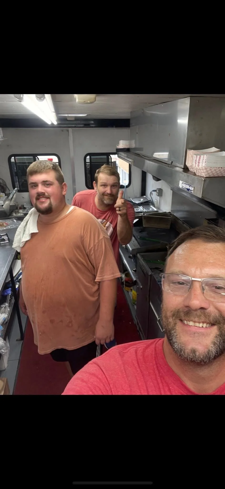 Three men inside a commercial kitchen, taking a selfie. Two men are smiling, and one man in the background is giving a thumbs-up. The kitchen has stainless steel appliances and counters.