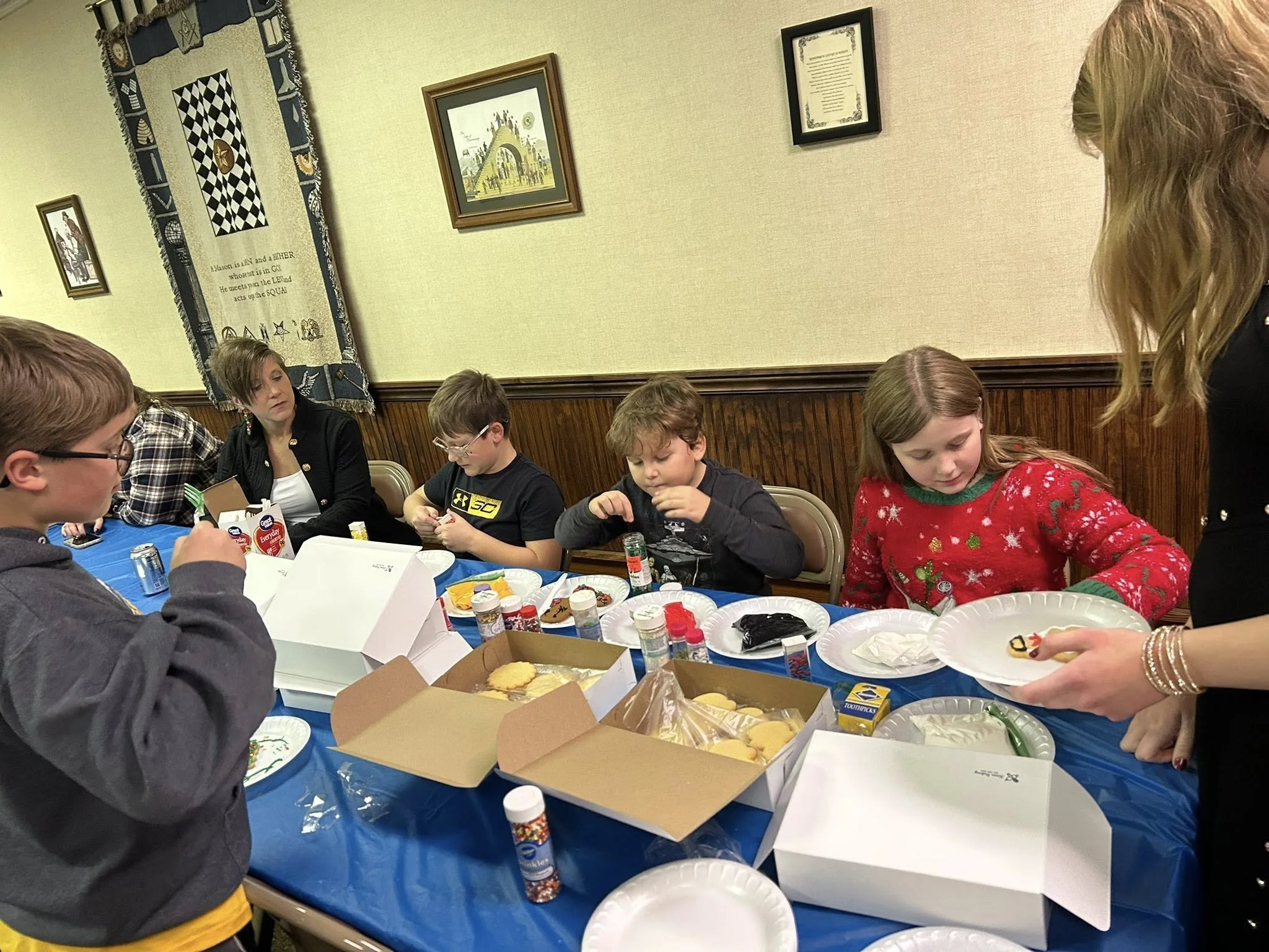 Children and adults gathered around a table with holiday cookies, icing, and decorating supplies, decorating cookies at a holiday party.