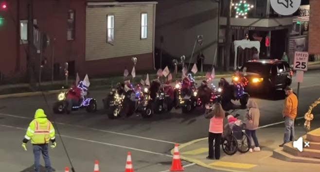 Sidewalk parade of decorated motorized scooters with American flags, surrounded by people, on a city street at night.