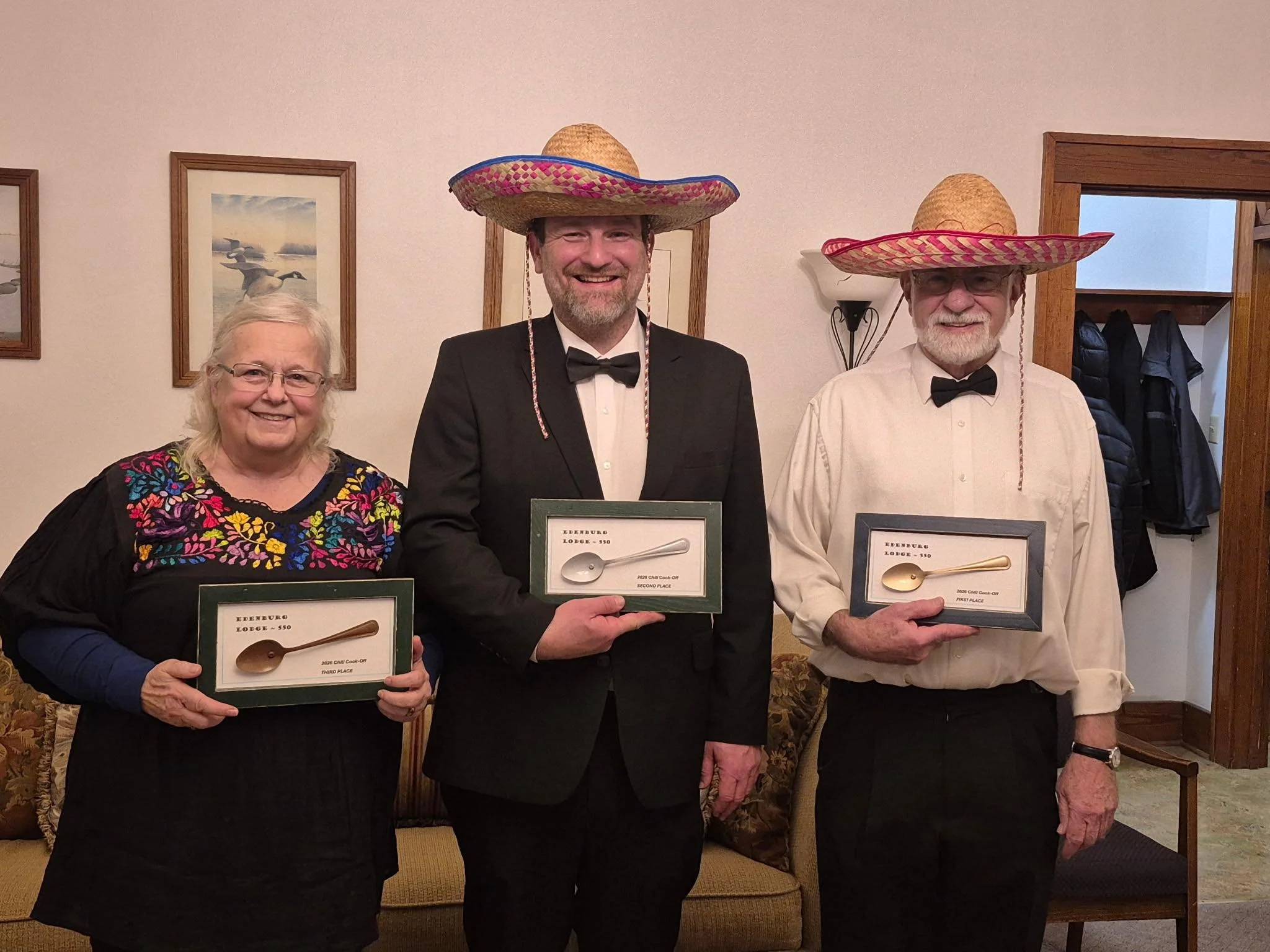 Three people standing together indoors, two men in tuxedos with sombreros and a woman in a black dress with colorful embroidery, each holding a framed award with a spoon image, at a lodge event.