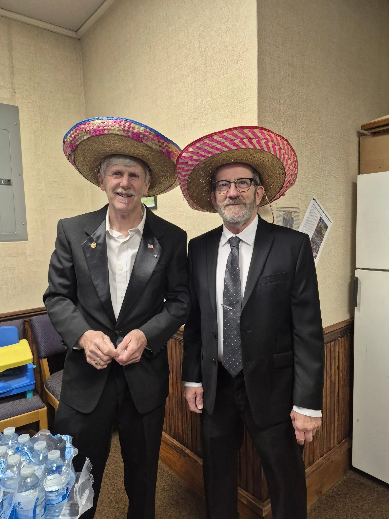 Two men in black suits and white shirts wearing large, colorful sombreros standing in a room with beige walls and wooden paneling. Water bottles are visible in the foreground.