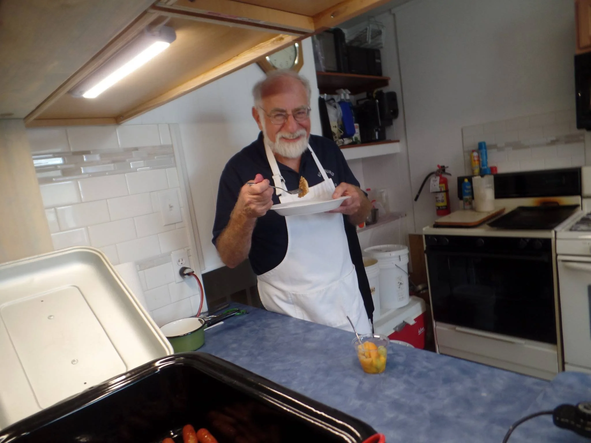 An elderly man with glasses and a white beard smiling while holding a spoon and a plate of food in a kitchen.