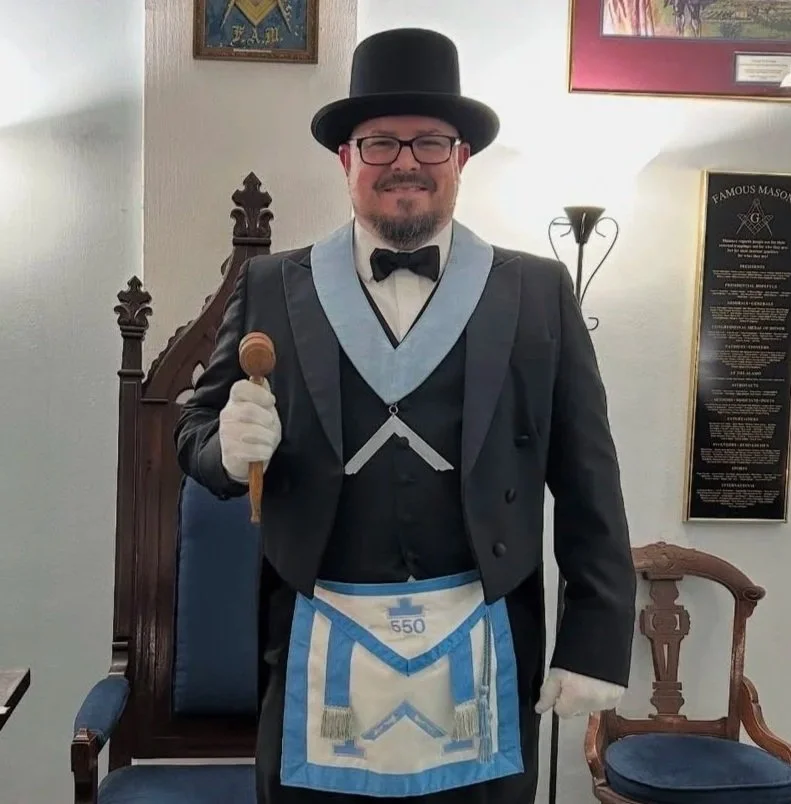 A man dressed in Masonic regalia, wearing a tuxedo with a top hat, bow tie, and glasses, holding a wooden gavel in his right hand, standing in a room with Masonic symbols and framed certificates on the wall.