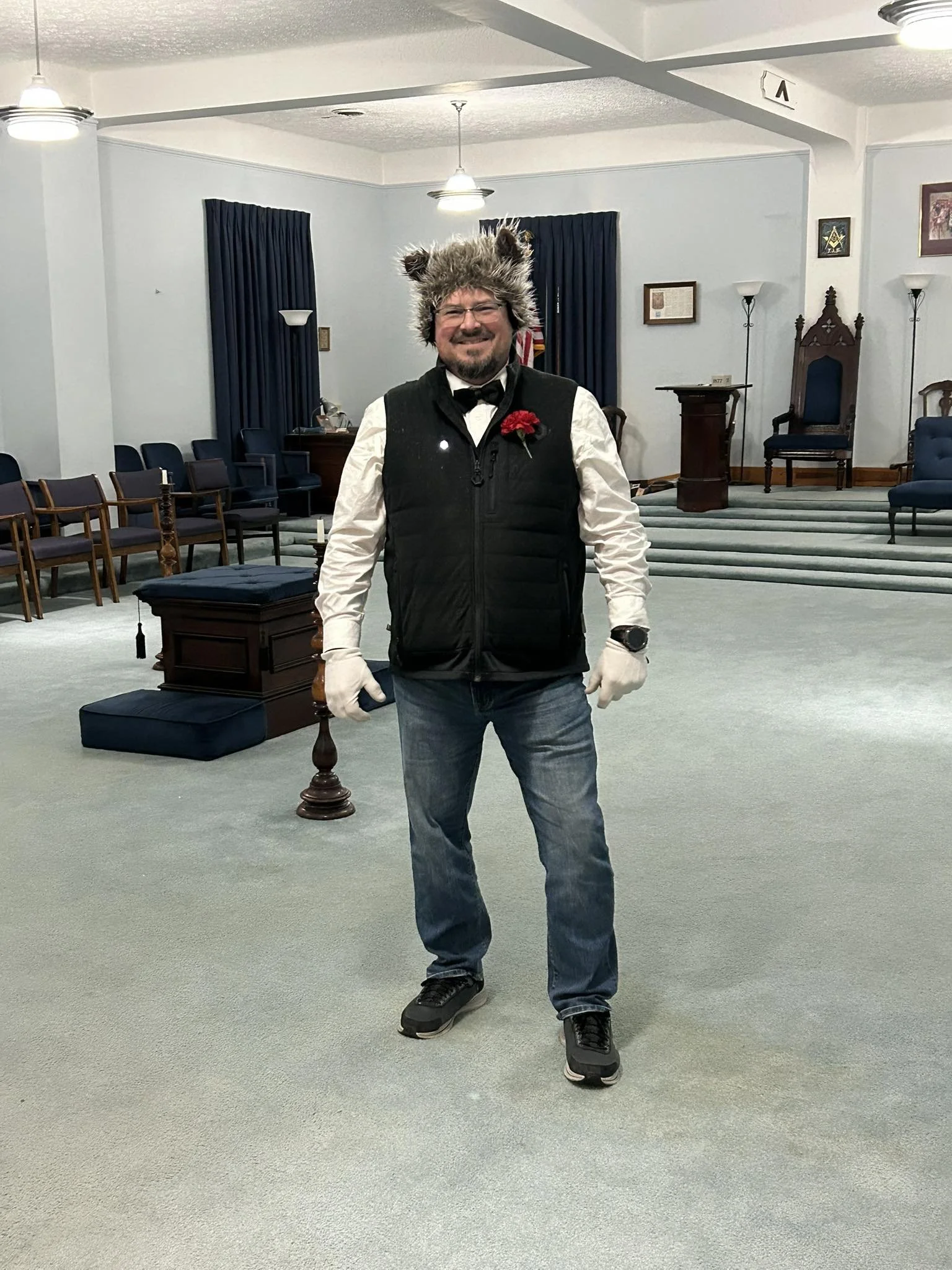 A man standing in a formal room dressed as a wolf, wearing a furry wolf hat, white shirt, black vest, jeans, gloves, and athletic shoes. He is smiling and wearing a red flower on his vest.