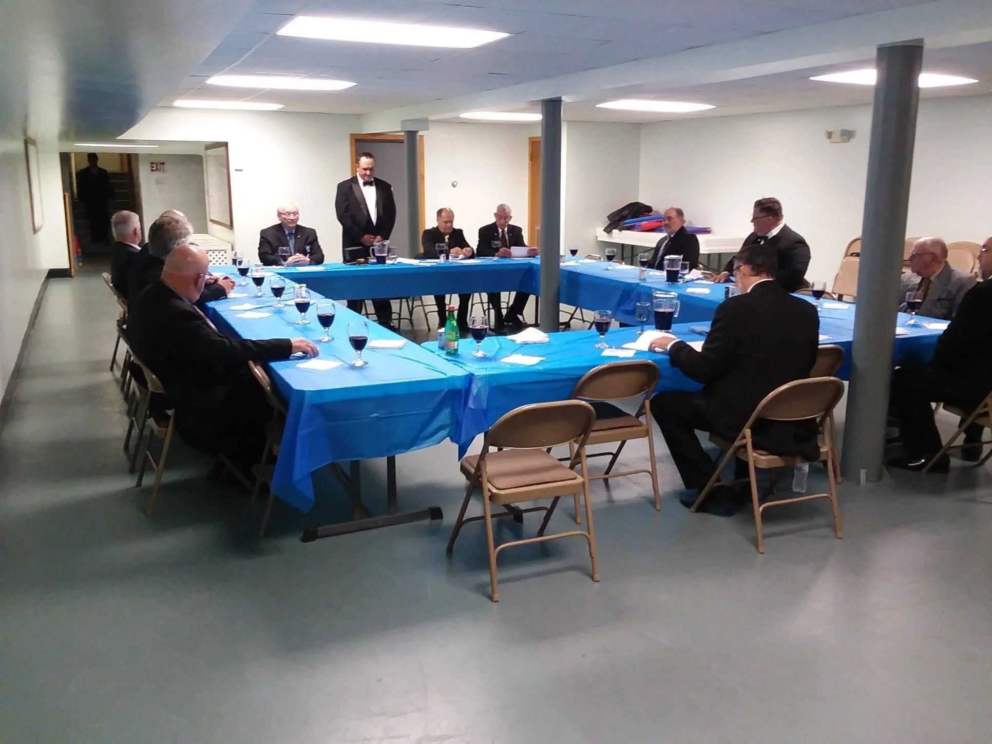A formal dinner or meeting setup with men in suits seated around a U-shaped table with blue tablecloths in a basement or conference room. One man in a tuxedo stands at the head of the table.