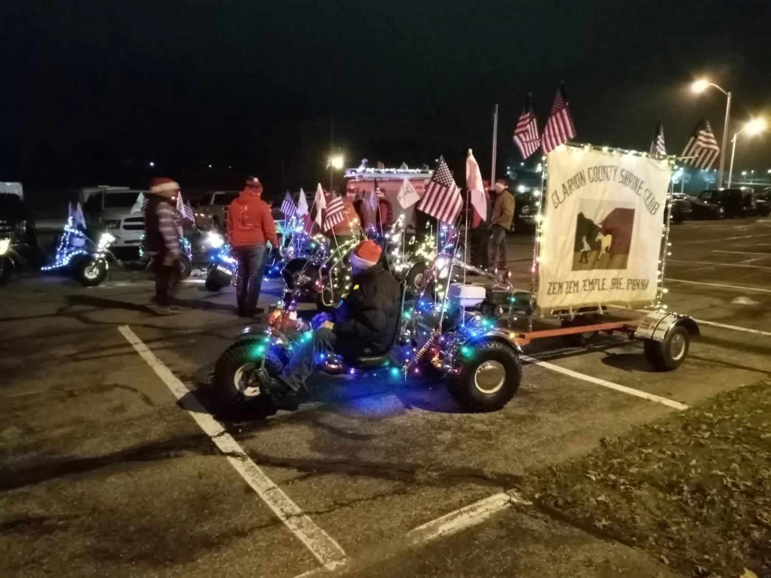 Nighttime parade scene with decorated tricycle and people dressed in warm clothing, displaying 'Glarion County Shrine Club' banner and American flags.