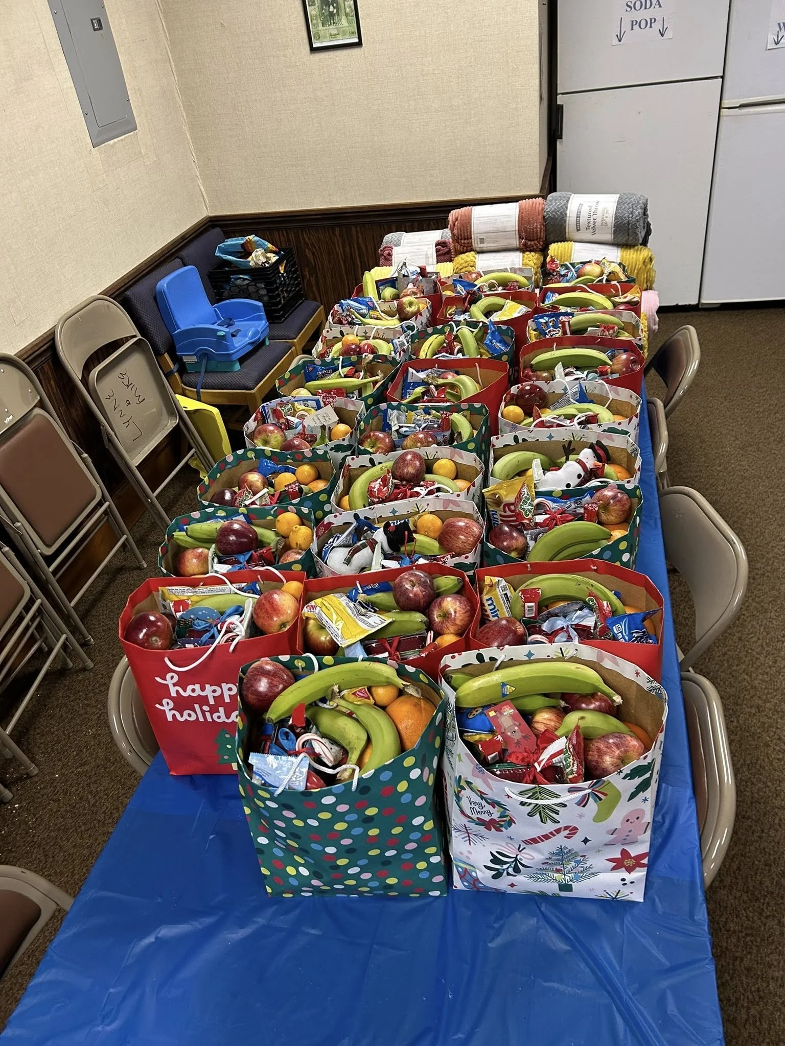 Table with holiday-themed gift bags filled with apples, bananas, oranges, and snacks, set in a room with chairs and folded towels in the background.